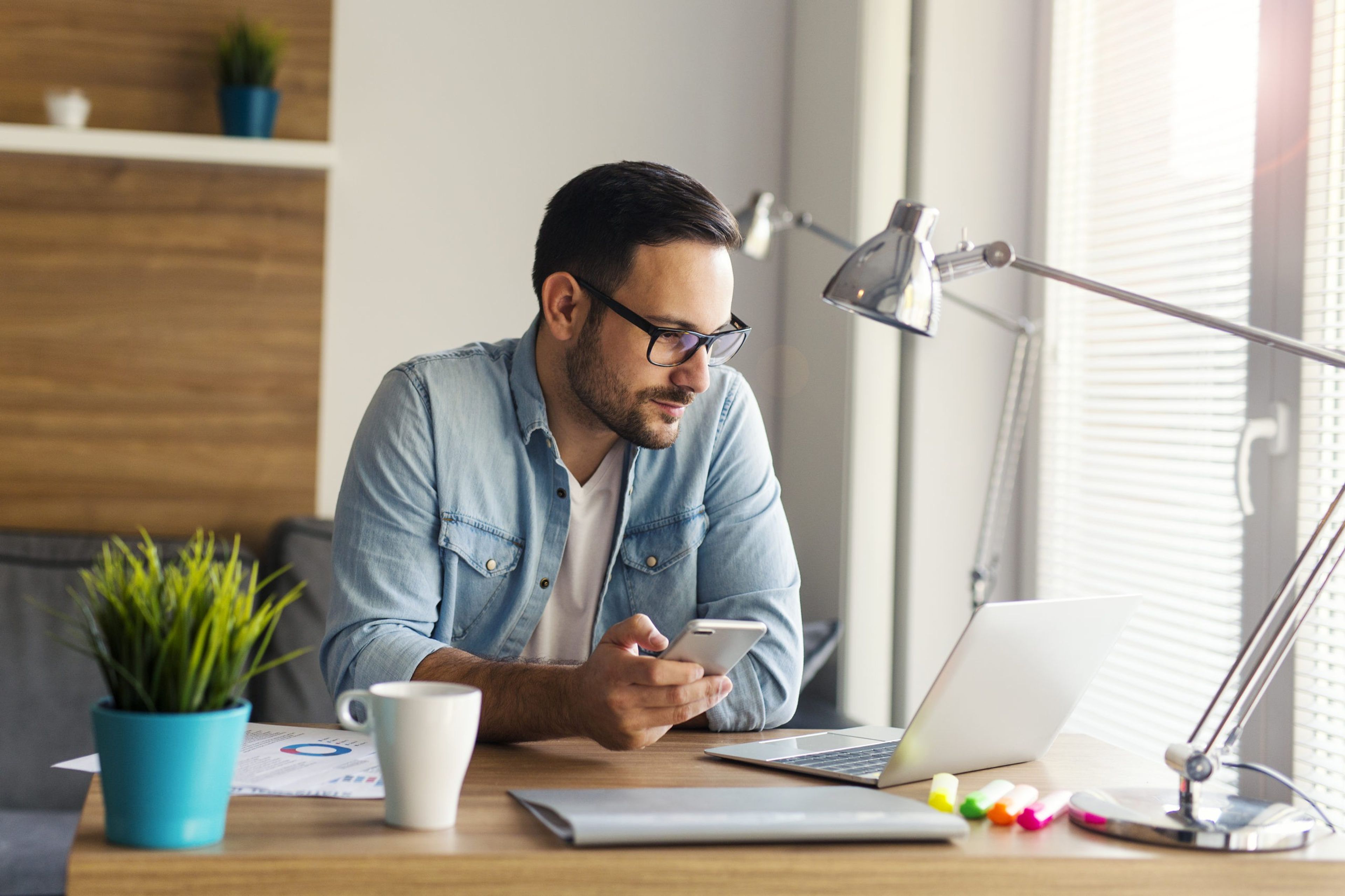 Man wearing collared shirt sitting at desk holding phone and looking at computer