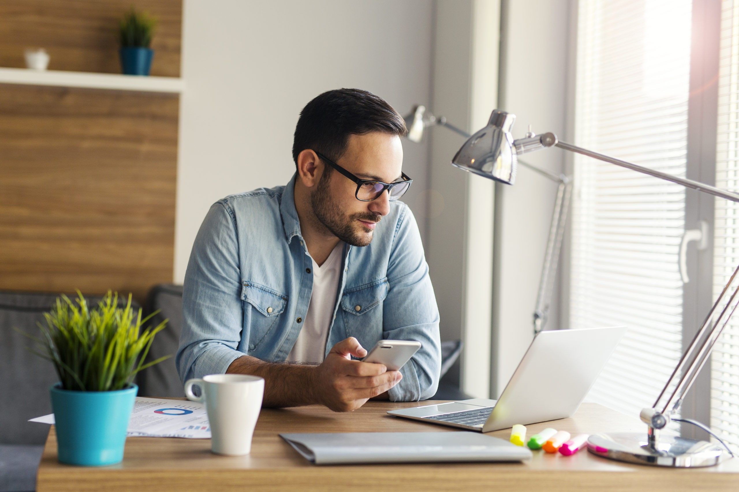 Man wearing collared shirt sitting at desk holding phone and looking at computer