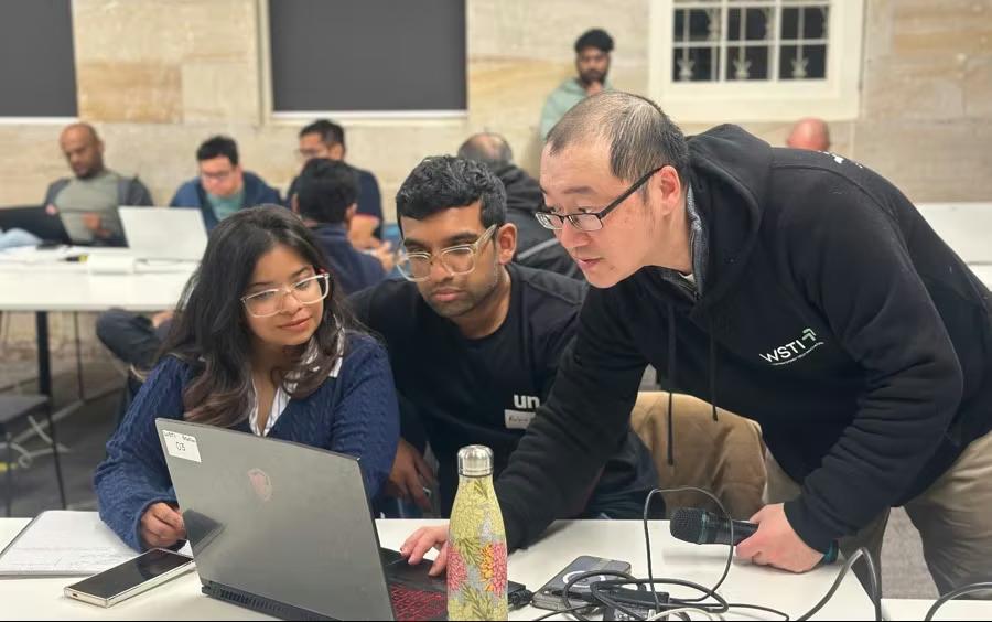 A WSTI mentor leaning over a laptop, working through a problem with two attendees at a workshop table.