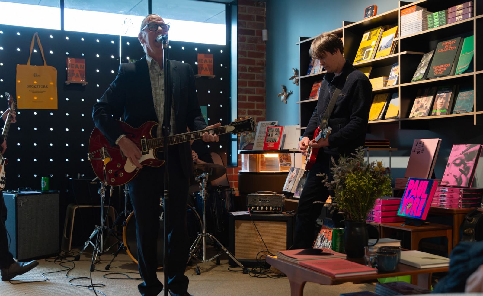 A live band performing inside Art Guide Bookstore in Northcote, two guitarists playing cherry red electric guitars before a twinkling dot-lit dark wall, surrounded by floor-to-ceiling shelves of colourful art publications, vintage timber furniture, and a signature yellow tote bag hanging in the background, the intimate space transformed into an electrifying live music venue with amplifiers and a drum kit tucked between the bookshelves.