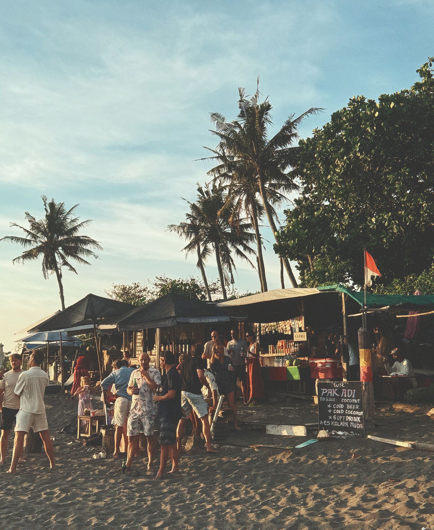 Sunset beach scene at Pererenan in Bali with casual beachfront warungs and bars, towering coconut palms, crowds gathering on sand, thatched roof structures, chalkboard menus, orange flags, and golden hour tropical atmosphere.