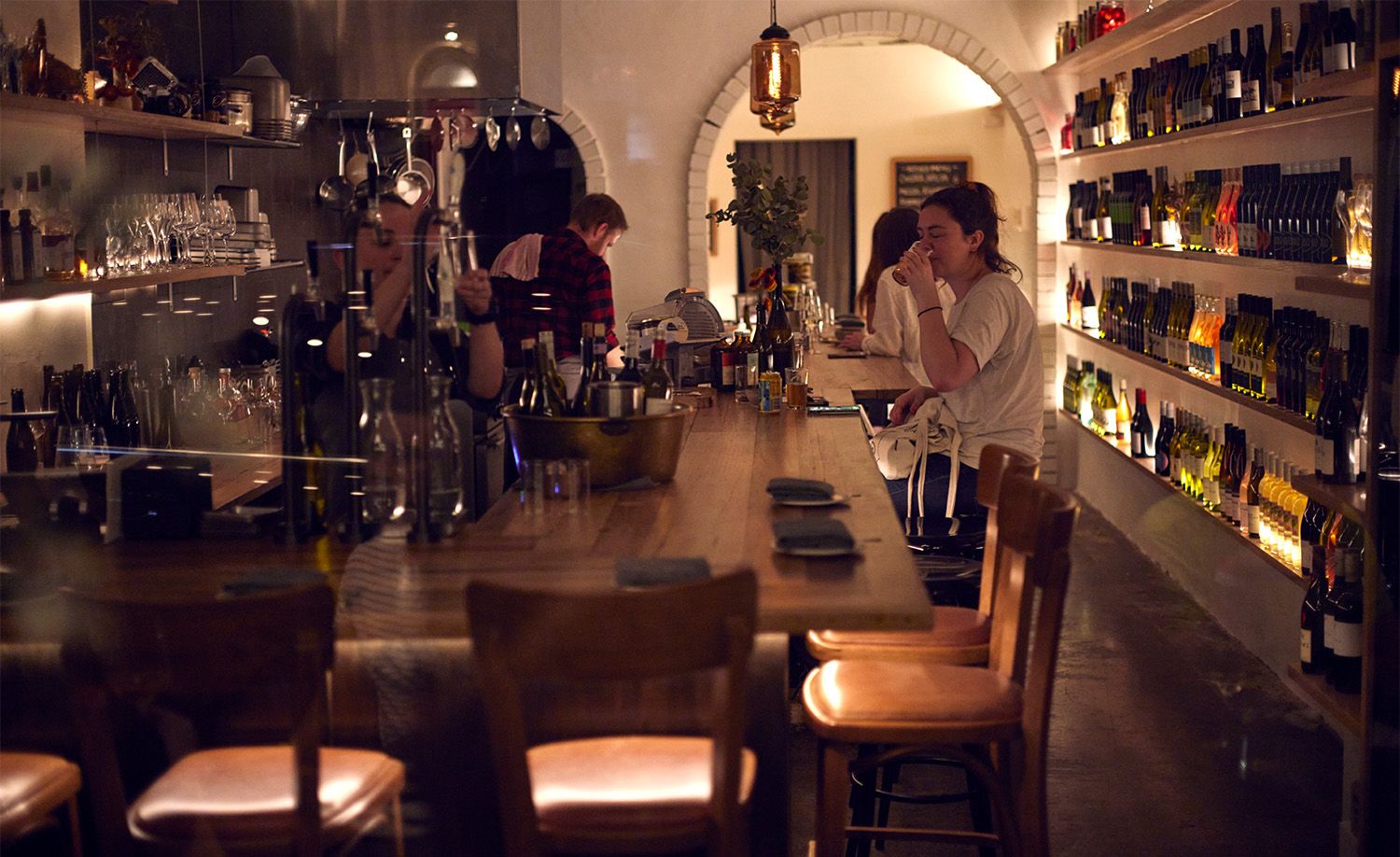 Wine bar Whitebark in North Melbourne featuring a long honey-toned timber communal bar stretching the full length of the narrow space, floor-to-ceiling wine bottle shelving lining the right-hand white brick wall bathed in warm amber underlighting, a white rendered arched brick ceiling drawing the eye to a glowing lantern pendant at the far end, a patron sipping wine at the bar while staff work behind the counter, creating the intimate and warmly lit atmosphere of a classic neighbourhood wine cellar bar.