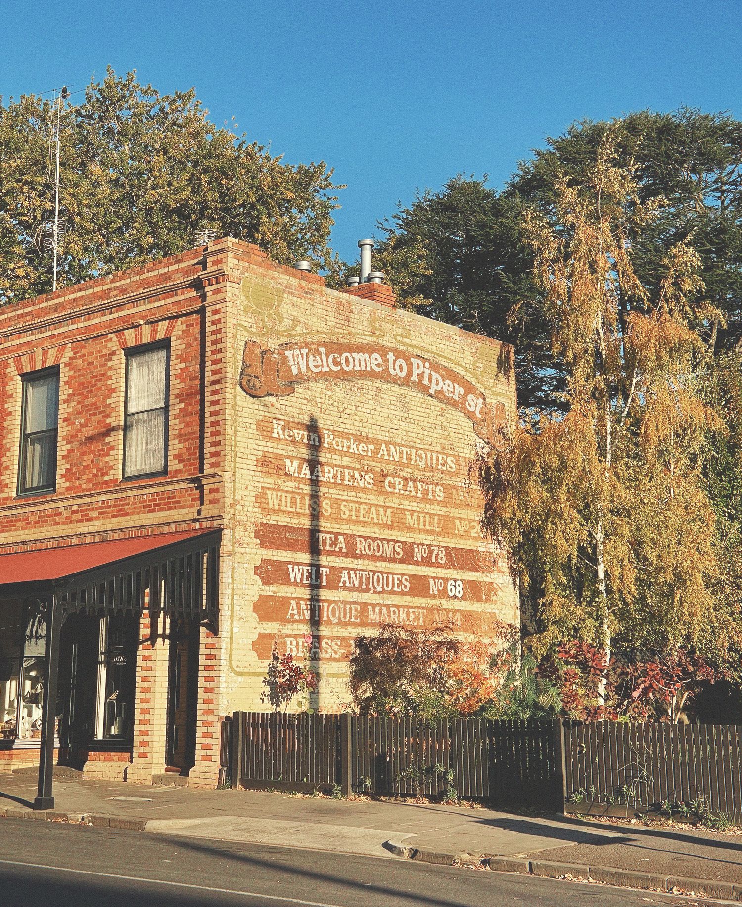 Heritage brick building on Piper Street in Kyneton with painted vintage signage advertising antiques, crafts and tea rooms, ornate parapet detailing, established trees with golden autumn foliage, timber picket fence, and warm afternoon sunlight illuminating country town streetscape.