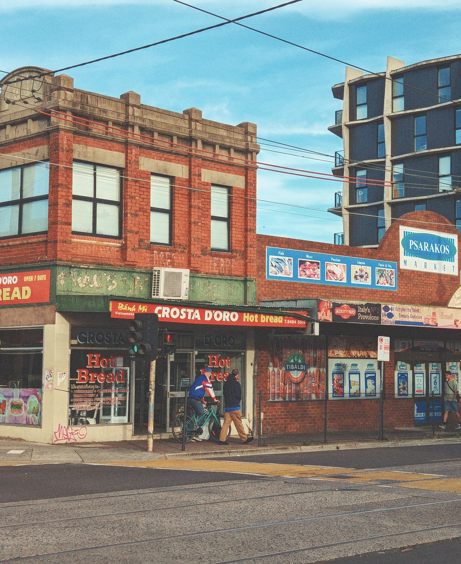High Street shopfronts in Thornbury with heritage red brick buildings featuring ornate parapet details, Crosta D'Oro bakery and neighbouring shops with bold signage, tram tracks in foreground, cyclist and pedestrian, modern apartment building rising behind, and inner-city Melbourne character.