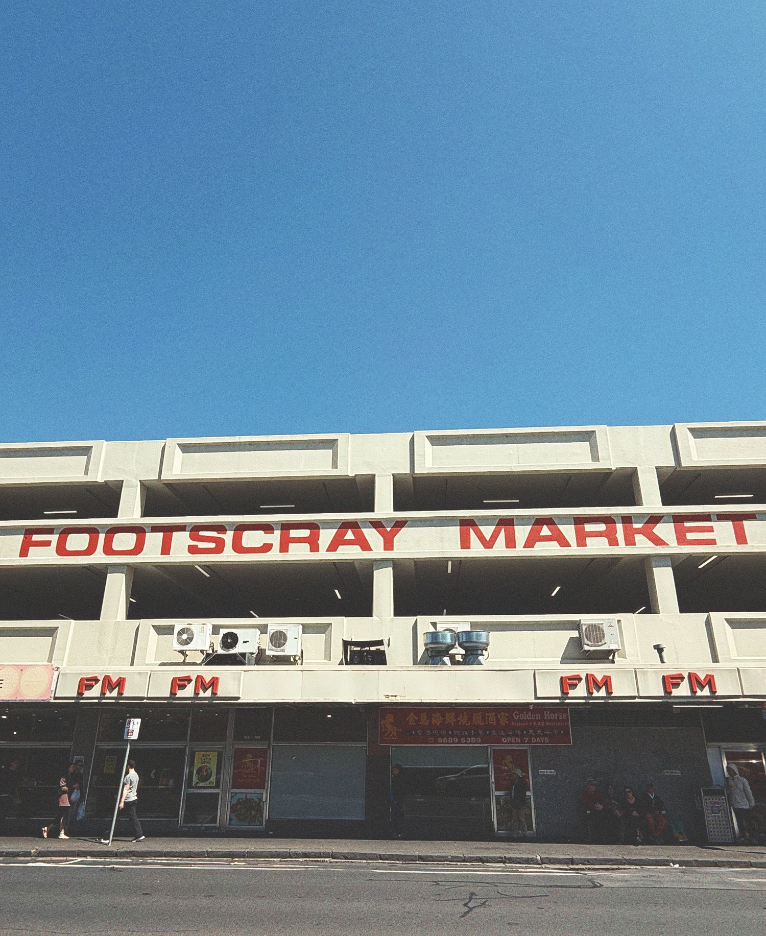 Footscray Market building facade with distinctive white brutalist architecture featuring geometric concrete balconies and openings, bold red signage spelling Footscray Market, ground floor shopfronts with FM lettering, air conditioning units visible, and clear blue sky above.