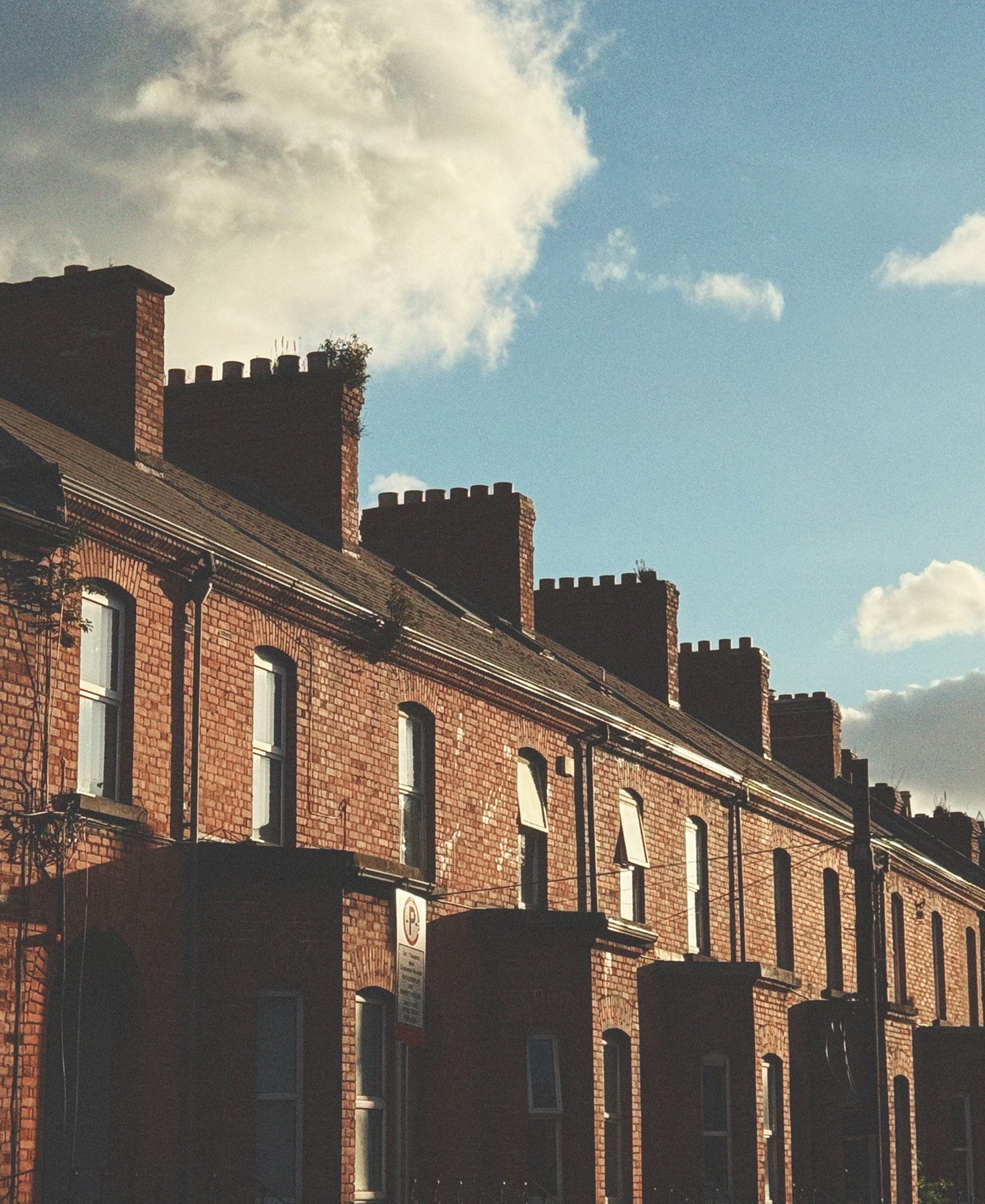 A residential street in Stoneybatter, Dublin featuring a long terrace of warm red brick Victorian workers' cottages with arched windows, their roofline punctuated by a rhythmic row of corbelled chimney stacks silhouetted dramatically against a vivid blue sky with billowing white clouds, the low afternoon sun casting deep shadows that accentuate the rich texture of the aged brickwork.