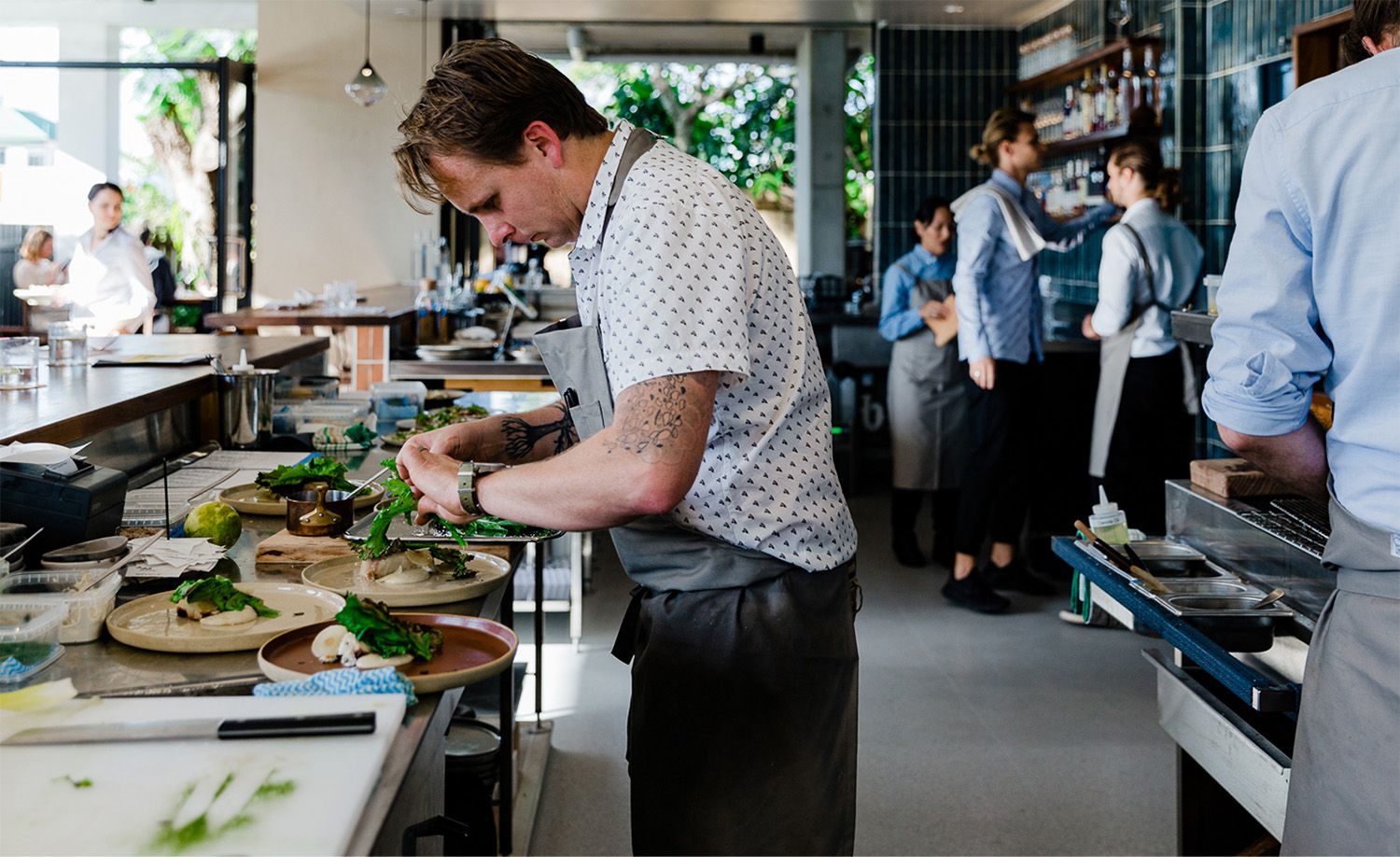 Chef Ben Devlin of Pipit restaurant focused intently over the pass as he carefully plates dishes with fresh green leaves, wearing a printed short-sleeve shirt and grey apron with a botanical tattoo visible on his forearm, front-of-house staff in blue shirts gathered in the background beside dark teal tiled walls and timber shelving, lush garden greenery visible through the open glass frontage behind, capturing the quiet concentration and considered craft at the heart of this acclaimed Northern Rivers restaurant.