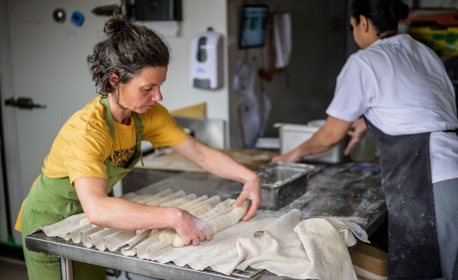 Baker Anna of Alex Bakes shaping baguette dough in the bakery kitchen, wearing a golden yellow t-shirt and sage green apron as she works a row of dough portions into a floured linen couche on a stainless steel bench, a second baker in a white t-shirt and dark apron working alongside her in the background, capturing the focused teamwork and quiet rhythm of an artisan bakery in full production.