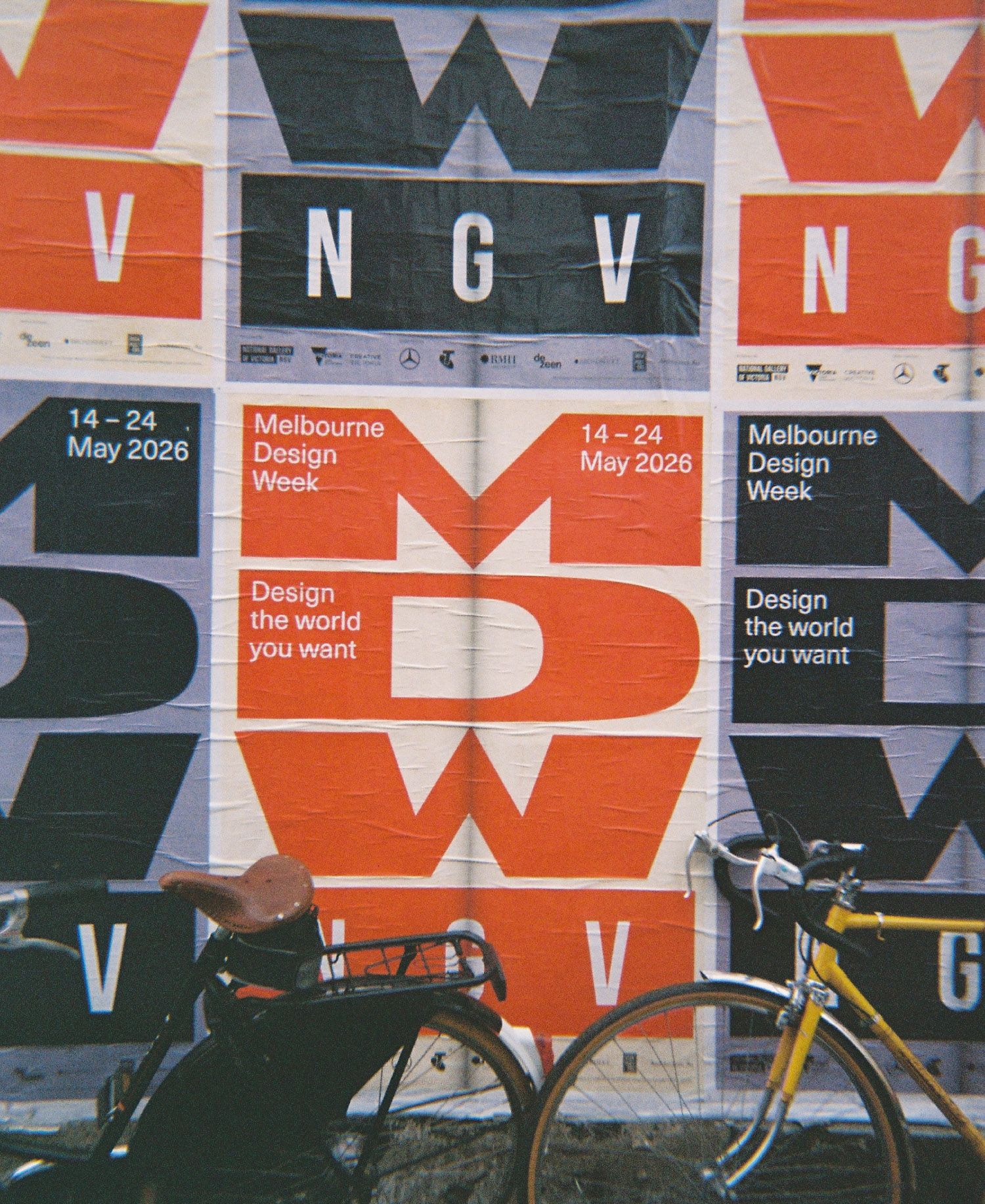Two bicycles — one black with a tan leather saddle, one mustard yellow — parked against a wall of wheat-pasted Melbourne Design Week 2026 posters featuring bold oversized MDW lettering in terracotta red, black and white, with NGV branding and the tagline "Design the world you want" repeated across the overlapping sheets, capturing the creative street energy of the Northside Design Ride.