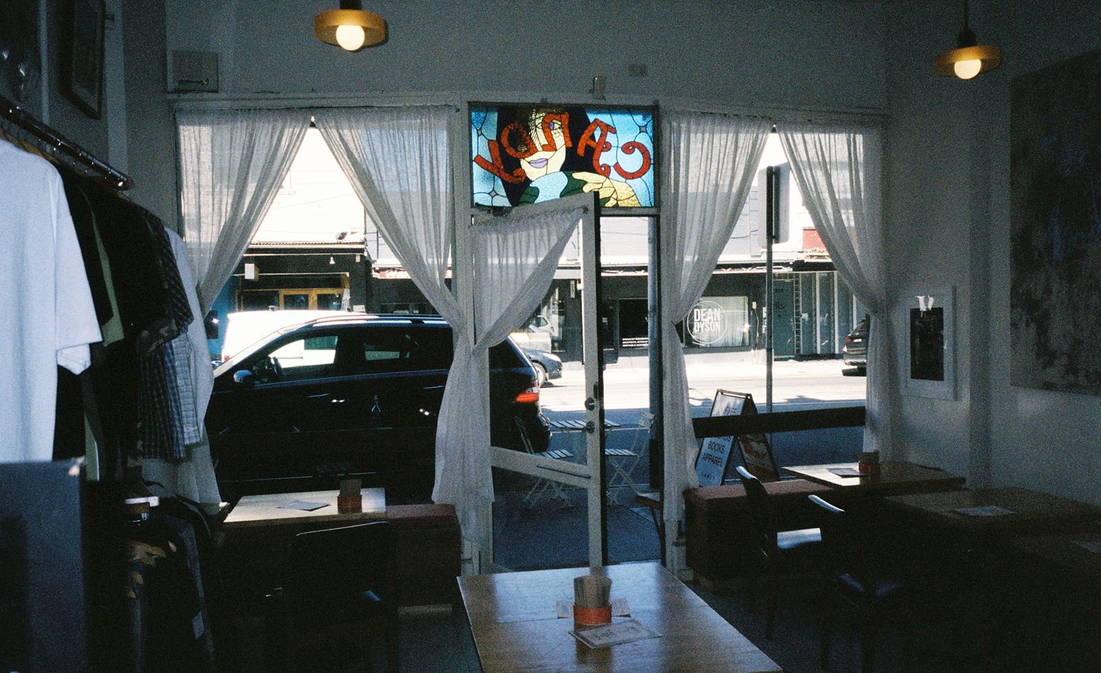 The interior of Candy in Melbourne looking out through the open shopfront door towards High Street, sheer white curtains framing large windows either side of the entrance, the vivid stained glass Candy panel above the door glowing with a colourful figurative motif in reverse, clothing racks, timber tables and dark bentwood chairs visible in the moody film-grain interior, the bright suburban streetscape of High Street and neighbouring shopfronts visible through the glass, capturing the relaxed and layered atmosphere of this characterful neighbourhood multi-space.