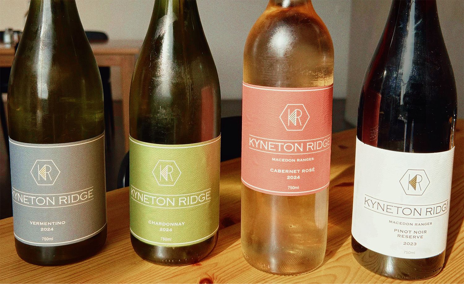 Four bottles of Kyneton Ridge Estate wine from the Macedon Ranges lined up on a blonde timber table, their elegantly designed labels in slate grey, olive green, terracotta pink and crisp white displaying the 2024 Vermentino, 2024 Chardonnay, 2024 Cabernet Rosé and 2023 Pinot Noir Reserve respectively, each bearing the geometric KR hexagon monogram, capturing the considered and proudly regional character of this acclaimed cool-climate Victorian wine estate.