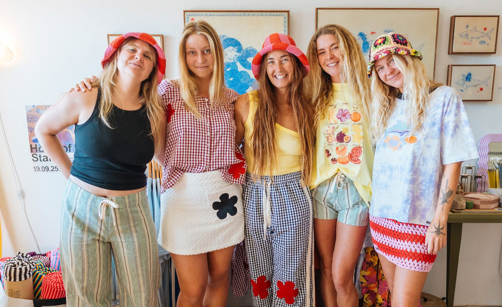 The team at Atmosea in Brunswick Heads including owner Maria Nilsson, five smiling women posing together in the store wearing an exuberant mix of gingham, linen, crochet bucket hats in coral and multicolour, a white textured mini skirt with floral appliqué, tie-dye and striped crochet shorts, framed botanical and marine illustrations visible on the white walls behind them, capturing the relaxed, playful and distinctly coastal spirit of the Atmosea team.