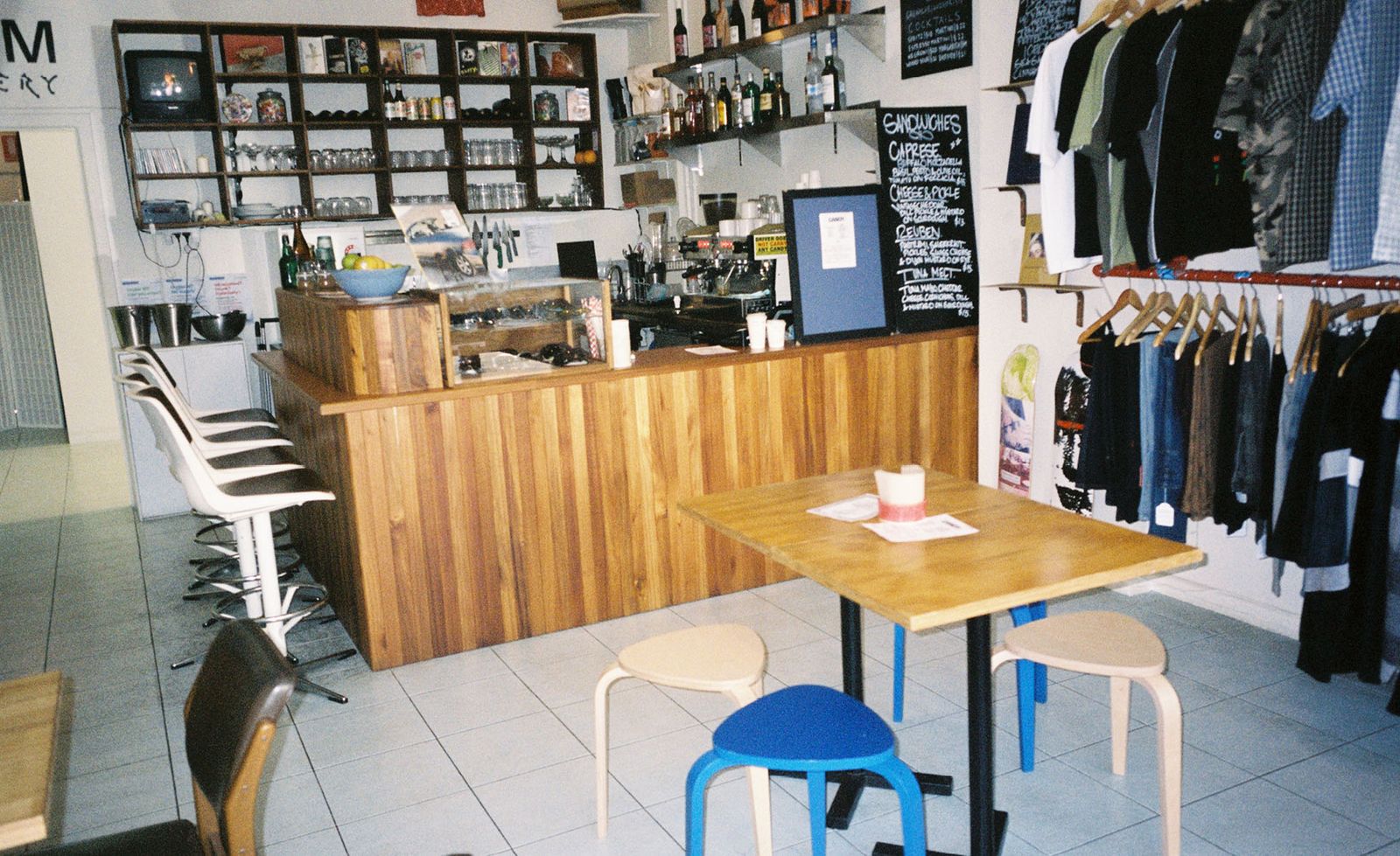 The interior of Candy in Melbourne revealing the full breadth of its multi-purpose character, a warm honey-toned timber bar counter at the centre flanked by mismatched seating including white swivel bar stools and cobalt blue Artek stools around a blonde timber café table, dark shelving above the bar housing a small television, records, glassware and spirits, handwritten sandwich and cocktail menus chalked on blackboards, and a clothing rack of garments lining the right-hand wall, the washed-out film grain of the photograph perfectly capturing the gloriously eclectic and unhurried spirit of this beloved neighbourhood haunt.