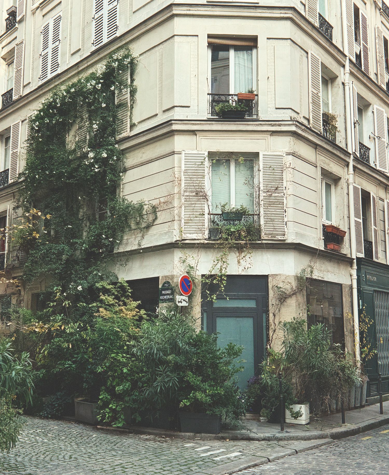 Corner Parisian building in Saint-Ambroise with cream rendered facade and traditional shutters, ivy and climbing plants cascading down walls, ground floor café or shop with lush potted greenery, weathered romantic character, cobblestone street, and quintessential Parisian neighbourhood atmosphere.