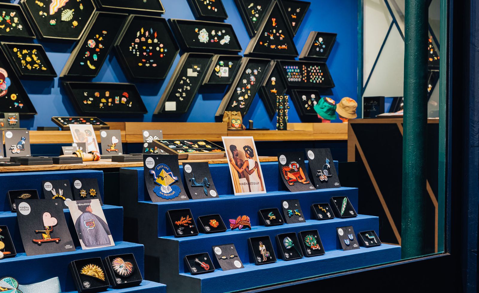 The interior of Macon et Lesquoy featuring tiered cobalt blue display steps showcasing individual boxed embroidered brooches in a dazzling array of motifs including animals, figures, and botanical forms, an entire cobalt blue wall covered floor to ceiling in hexagonal and diamond black velvet display frames presenting the full breadth of the collection, a campaign postcard and branded packaging adding warmth to the blonde timber counter behind, creating an exuberantly colourful and meticulously curated retail environment that feels as much cabinet of curiosities as it does boutique.