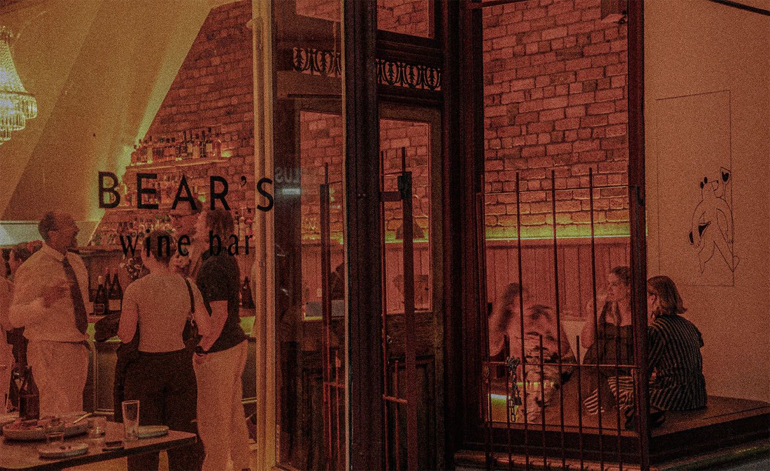 Wine bar Bear's Wine Bar in North Melbourne photographed from the street on a warm evening, the amber-lit exposed brick interior glowing through large shopfront windows with the bar name etched on the glass, patrons mingling at the bar stocked with backlit bottles while others are seated in an intimate wrought iron window booth beside a minimal line art print, a crystal chandelier visible in the upper left corner, the whole scene bathed in a deep red-amber warmth that captures the seductive and inviting atmosphere of this neighbourhood wine bar at its best.