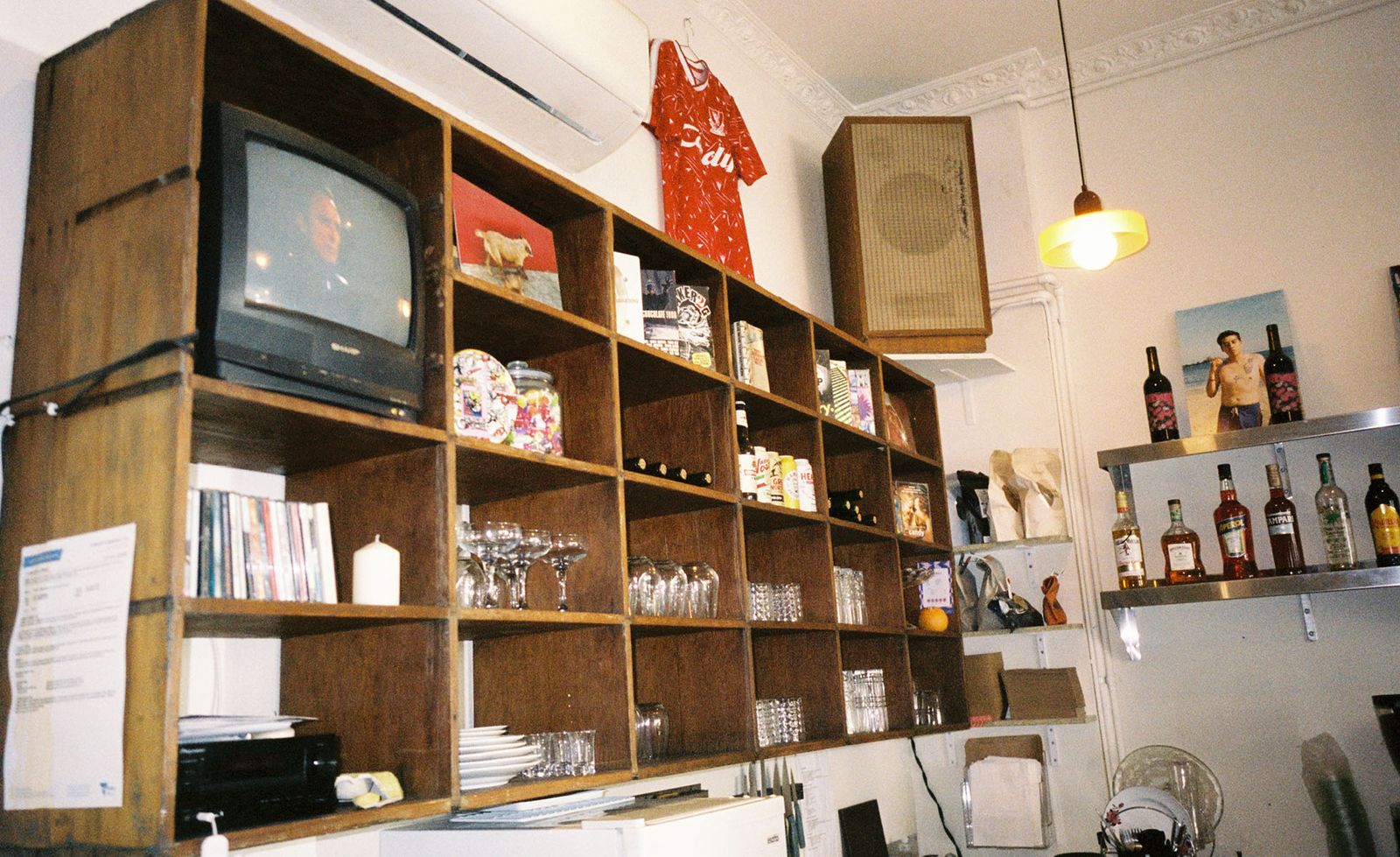 A wall of dark timber cubby shelving inside Candy in Melbourne housing an eclectic collection of glassware, vinyl records, zines, a sticker-covered jar, white pillar candles and miscellaneous curiosities, a vintage Sharp CRT television playing in the top corner, a red football jersey hanging above a large timber speaker box mounted on the wall, spirits bottles and a ceramic bust visible on steel shelving to the right beneath an ornate plaster cornice ceiling, a warm pendant light casting a golden glow across the scene, captured in film grain that perfectly suits the nostalgic and gloriously cluttered atmosphere of this one-of-a-kind Melbourne local.