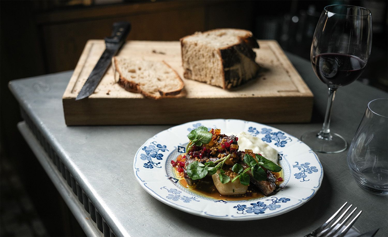 A beautifully composed dish at Restaurant Janine in Batignolles, Paris, served on a blue and white floral porcelain plate atop a stainless steel pass, a carefully plated combination of seared meat, soft potato, whipped cream, pomegranate seeds, mustard and fresh watercress leaves in a golden sauce, a timber bread board with sliced sourdough and a dark-handled knife alongside a glass of deep red wine completing the scene, capturing the intimate and considered atmosphere of a classic Parisian neighbourhood bistro.