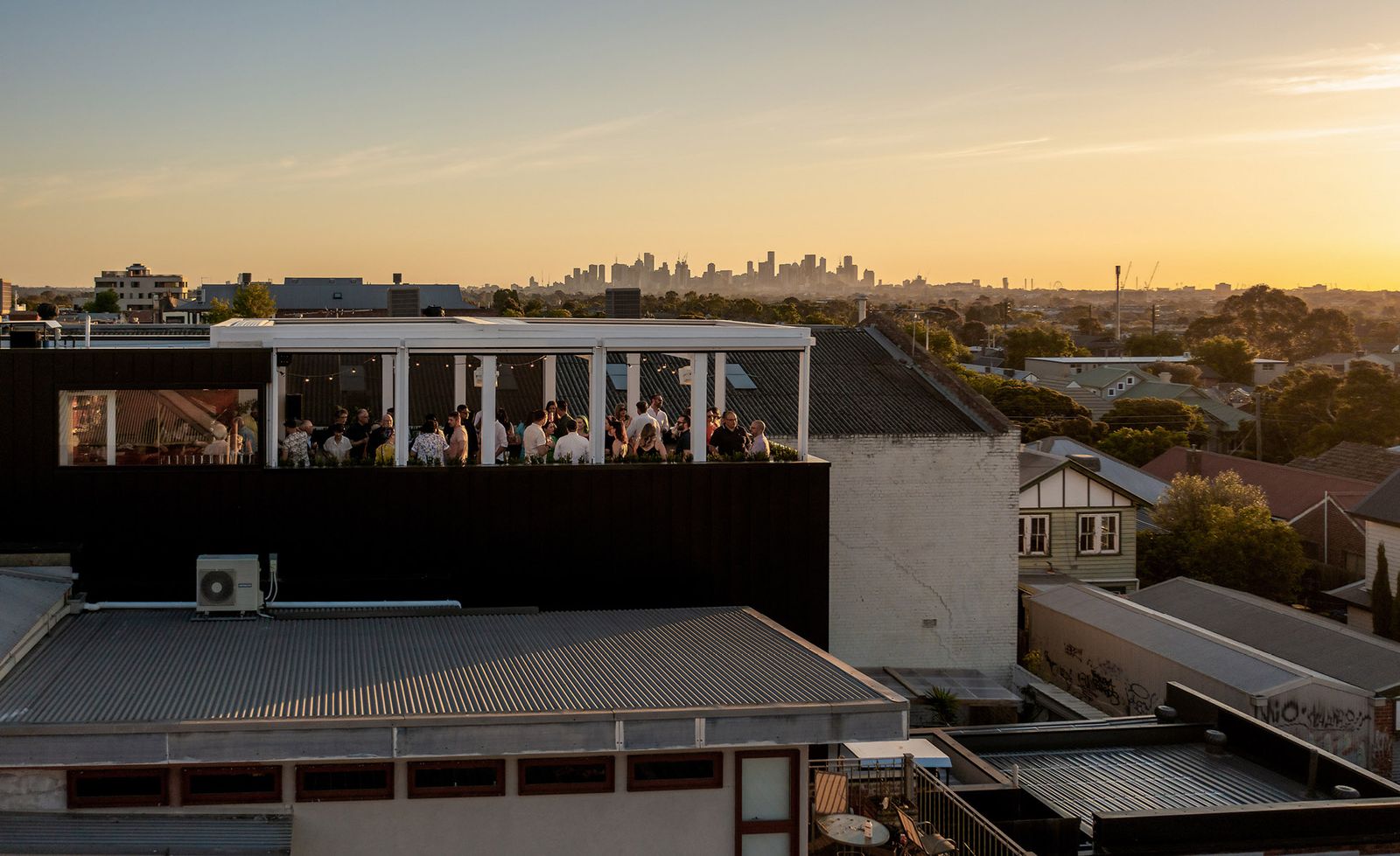 An aerial view of Gigi Rooftop bar in Thornbury at golden hour, a crowd of silhouetted patrons gathered beneath a white pergola structure strung with festoon lighting on top of a dark commercial building, the vast Melbourne suburban roofscape of corrugated iron, weatherboard cottages and mature gum trees stretching to the horizon where the city skyline rises as a hazy silhouette against a glowing amber and golden sunset sky, perfectly capturing why this rooftop has become one of Melbourne's most treasured evening destinations.