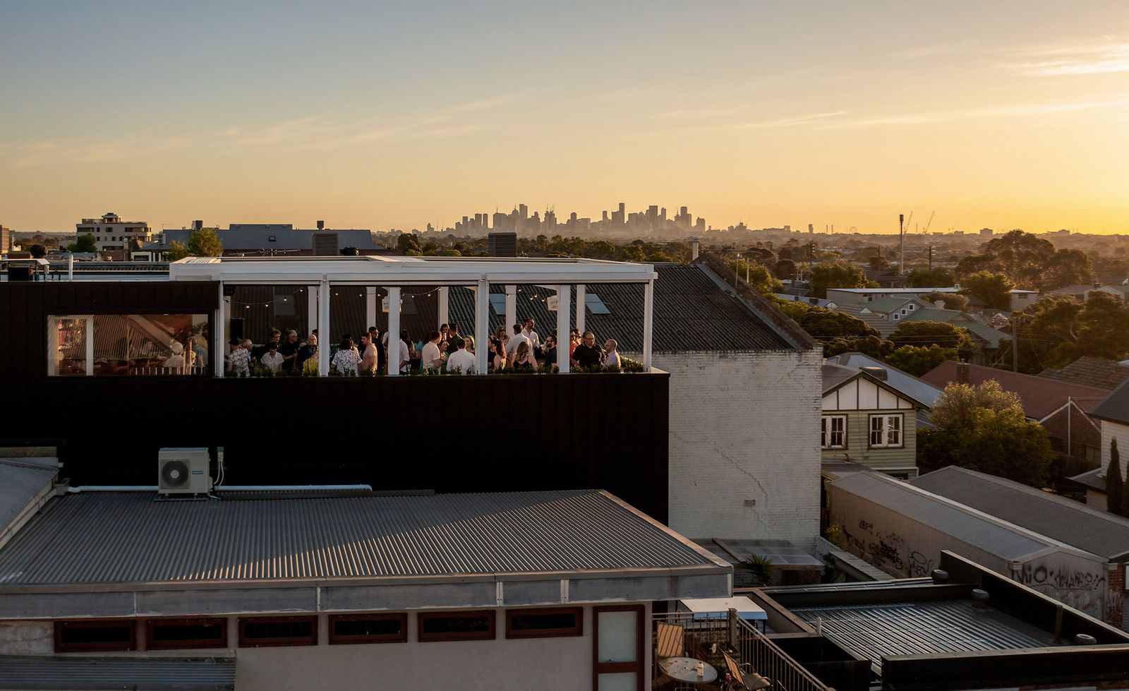 An aerial view of Gigi Rooftop bar in Thornbury at golden hour, a crowd of silhouetted patrons gathered beneath a white pergola structure strung with festoon lighting on top of a dark commercial building, the vast Melbourne suburban roofscape of corrugated iron, weatherboard cottages and mature gum trees stretching to the horizon where the city skyline rises as a hazy silhouette against a glowing amber and golden sunset sky, perfectly capturing why this rooftop has become one of Melbourne's most treasured evening destinations.