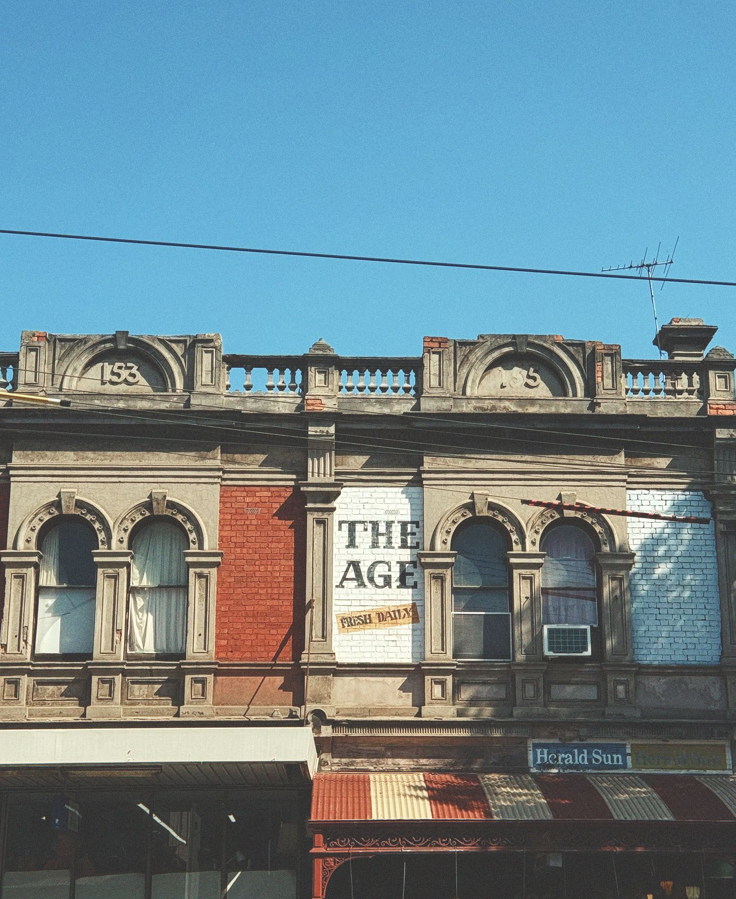Heritage Victorian-era shopfronts on Errol Street in North Melbourne with ornate rendered facades featuring arched windows and decorative parapet with 1953 date markers, vintage The Age newspaper signage, mixed red brick and painted sections, corrugated iron awnings, TV aerial, and clear blue sky.