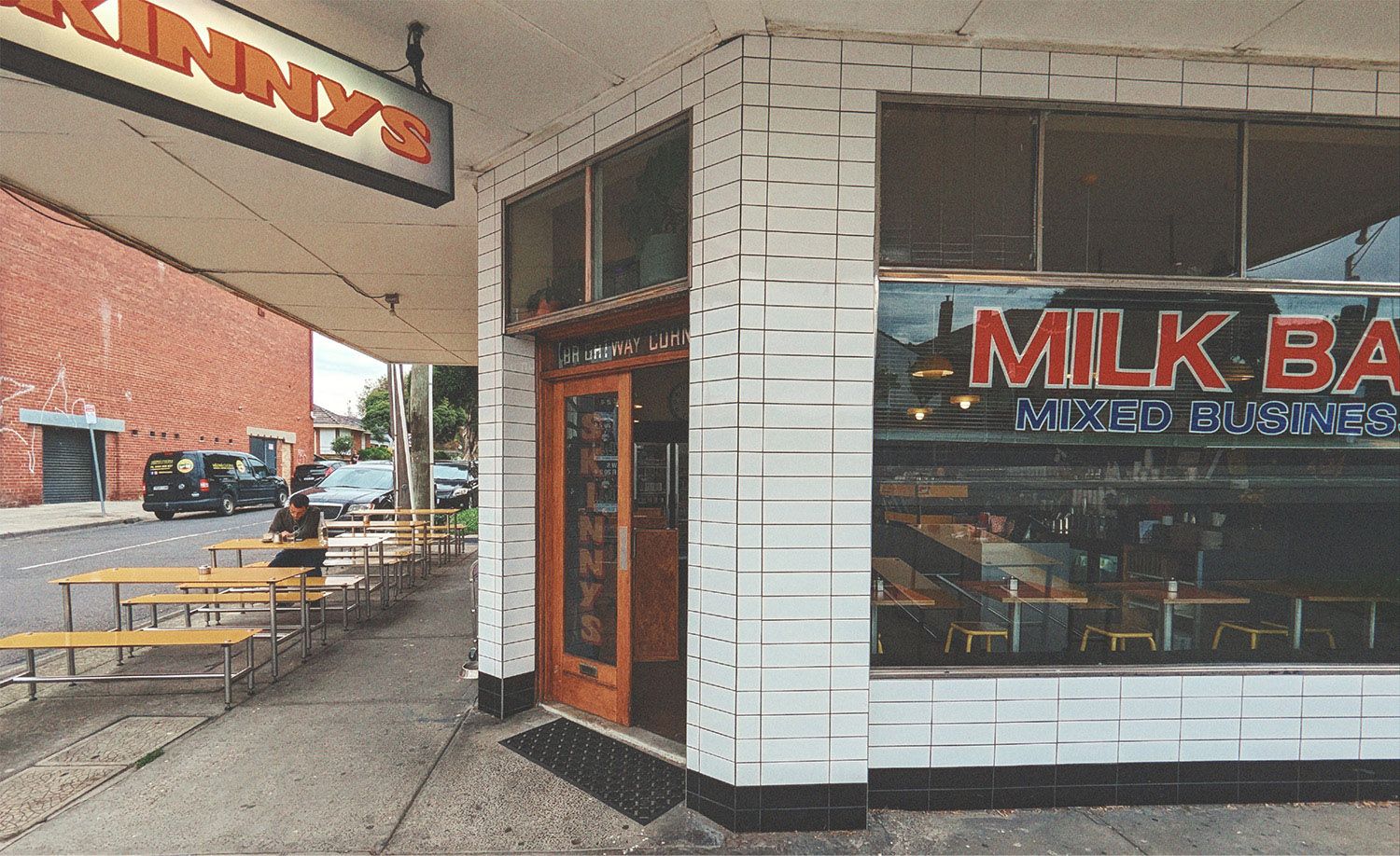Diner Skinnys in Thornbury occupying a classic corner milk bar premises with white subway-tiled walls and bold red and blue Milk Bar Mixed Business signage on the shopfront window, a warm timber door with the Skinnys name etched in glass, mustard yellow picnic bench seating lining the footpath beneath a covered awning where a solitary patron sits with a coffee, the retro illuminated Skinnys sign projecting above the entrance, capturing the nostalgic and unpretentious charm of a beloved neighbourhood diner paying affectionate homage to the great Australian milk bar.