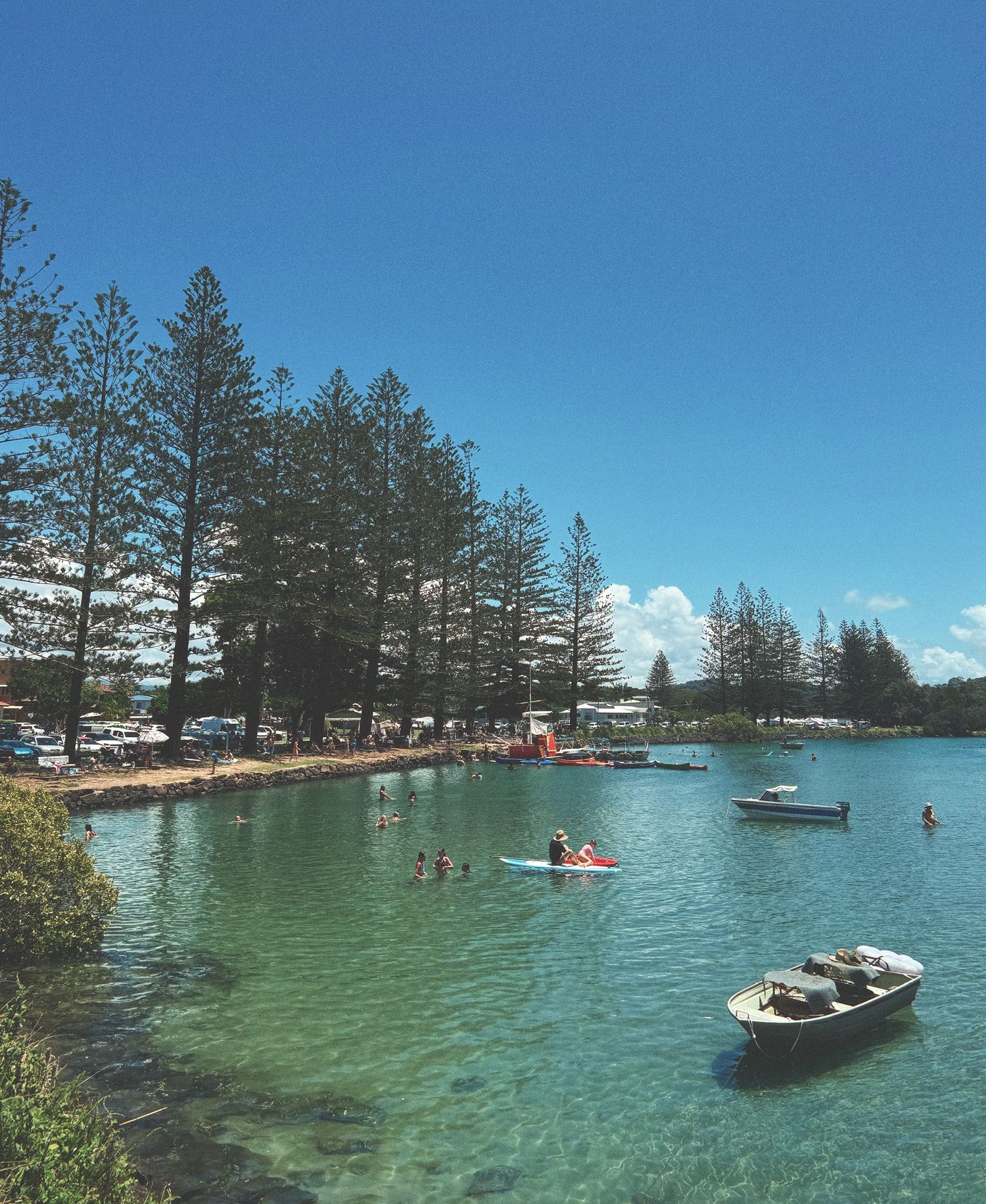 Brunswick River swimming spot in Brunswick Heads with crystal clear turquoise water, swimmers and kayakers enjoying calm estuary, small boats moored, towering Norfolk pines lining sandy shore, families relaxing under tree shade, and bright blue summer sky.