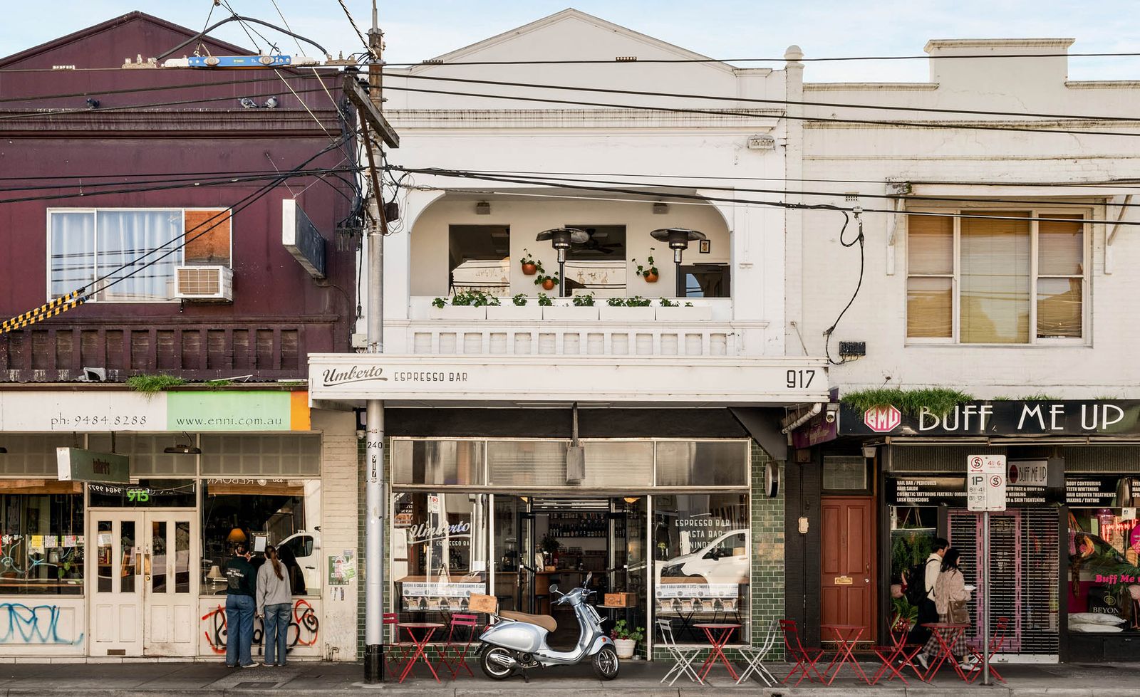 High Street Thornbury centred on Umberto Espresso Bar at number 917, its white-rendered two-storey Victorian facade with ornate arched first-floor balcony dressed with terracotta pots and trailing greenery above a dark green tiled ground floor shopfront, red bistro tables and chairs spilling onto the footpath beside a silver Vespa scooter, the eclectic neighbouring shopfronts of a typical inner-north Melbourne high street stretching either side beneath a pale blue sky, capturing the relaxed Italian character of this cherished neighbourhood institution with Gigi Rooftop occupying the floor above.