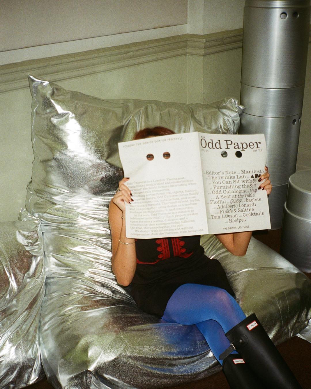 Person reading Ödd Paper through eye holes, seated on a shiny silver chair, wearing blue tights and black boots.