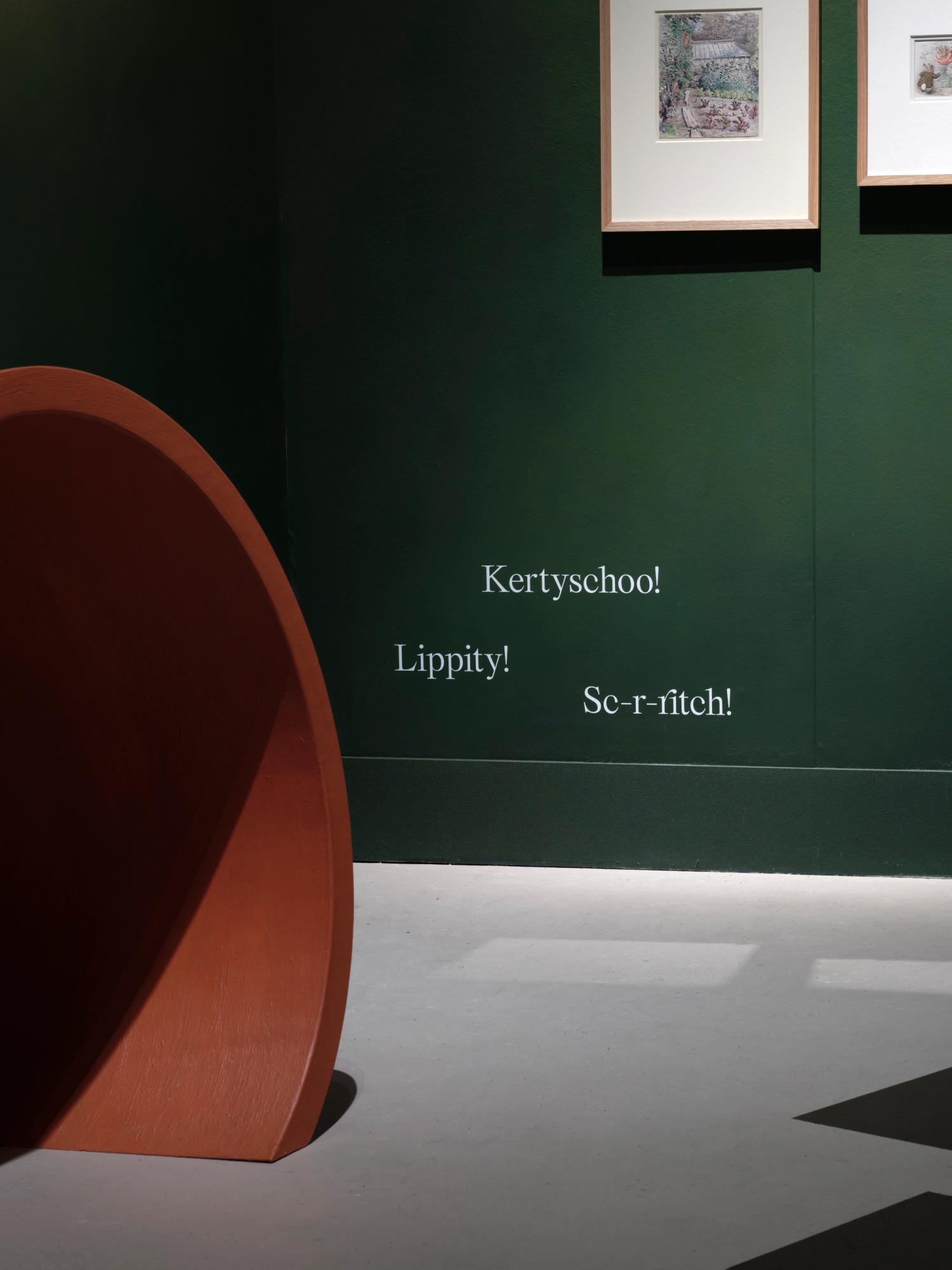 A photograph of a gallery space with a large rust-coloured curved tunnel structure in the foreground and a green wall behind with onomatopoeic text printed in white on the wall