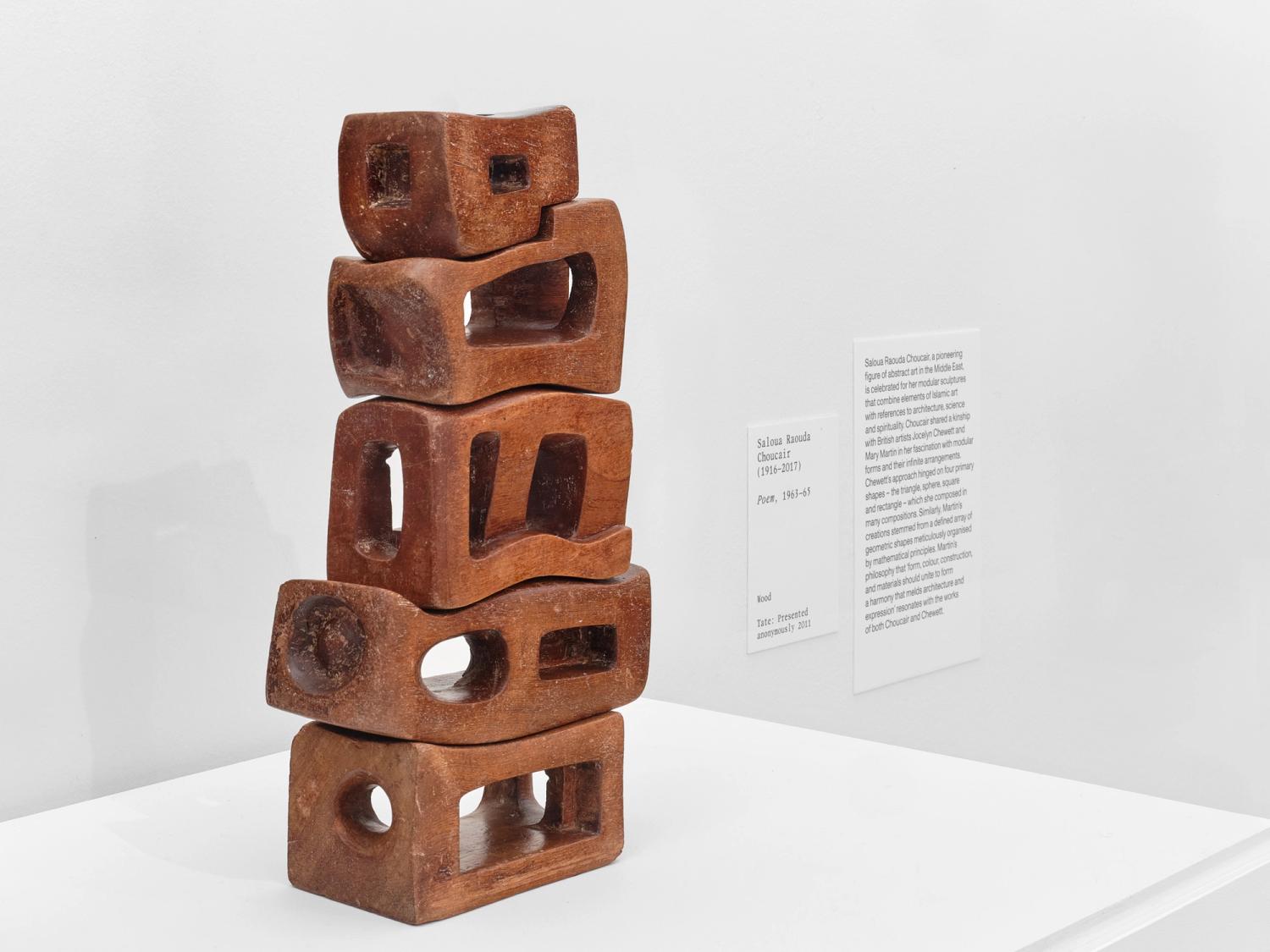 From the exhibition 'Beyond Form', the artwork 'Poem' from 1963-65 from the artist Saloua Raouda Choucair. The artworks is of 5 brown carved wooden shapes that resemble bricks stacked on top of one another. In the background is a white object label