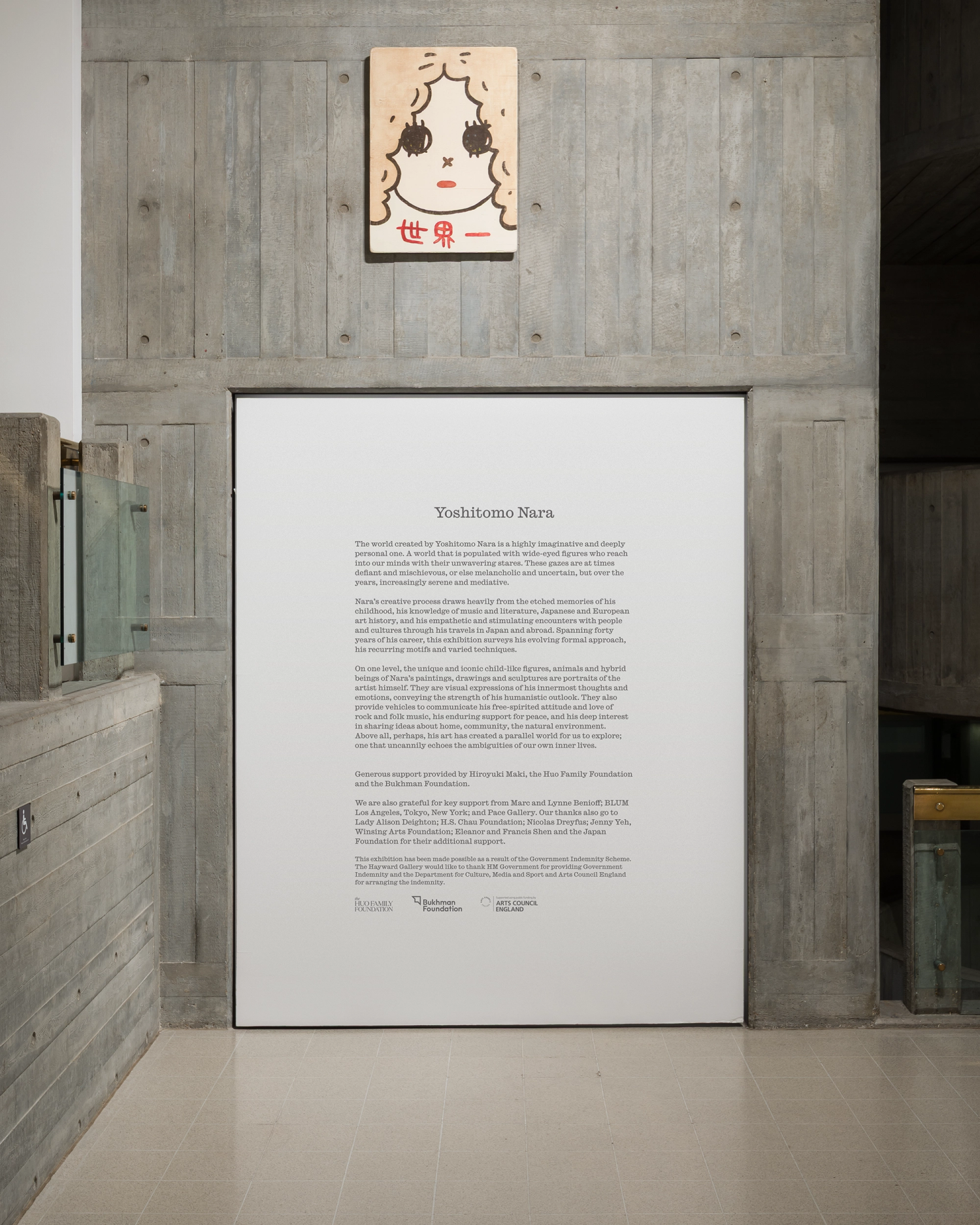 A white wall panel with black text sits inset into a textured concrete wall in a gallery. An artwork with Japanese script sits above.