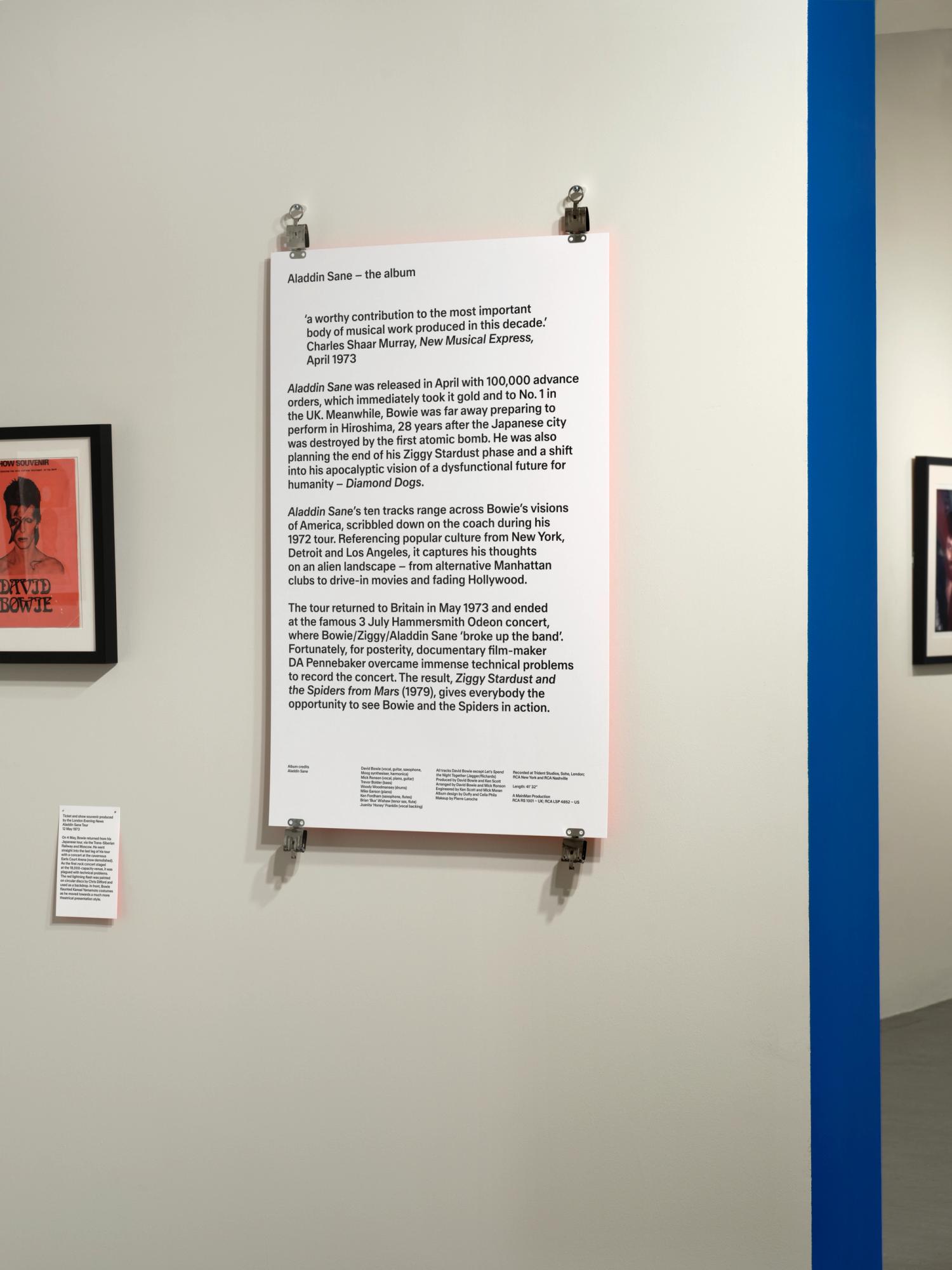 A text panel in a gallery space. The panel has a red glow reflecting against the white wall. It is secured with stainless steel clips at each corner.