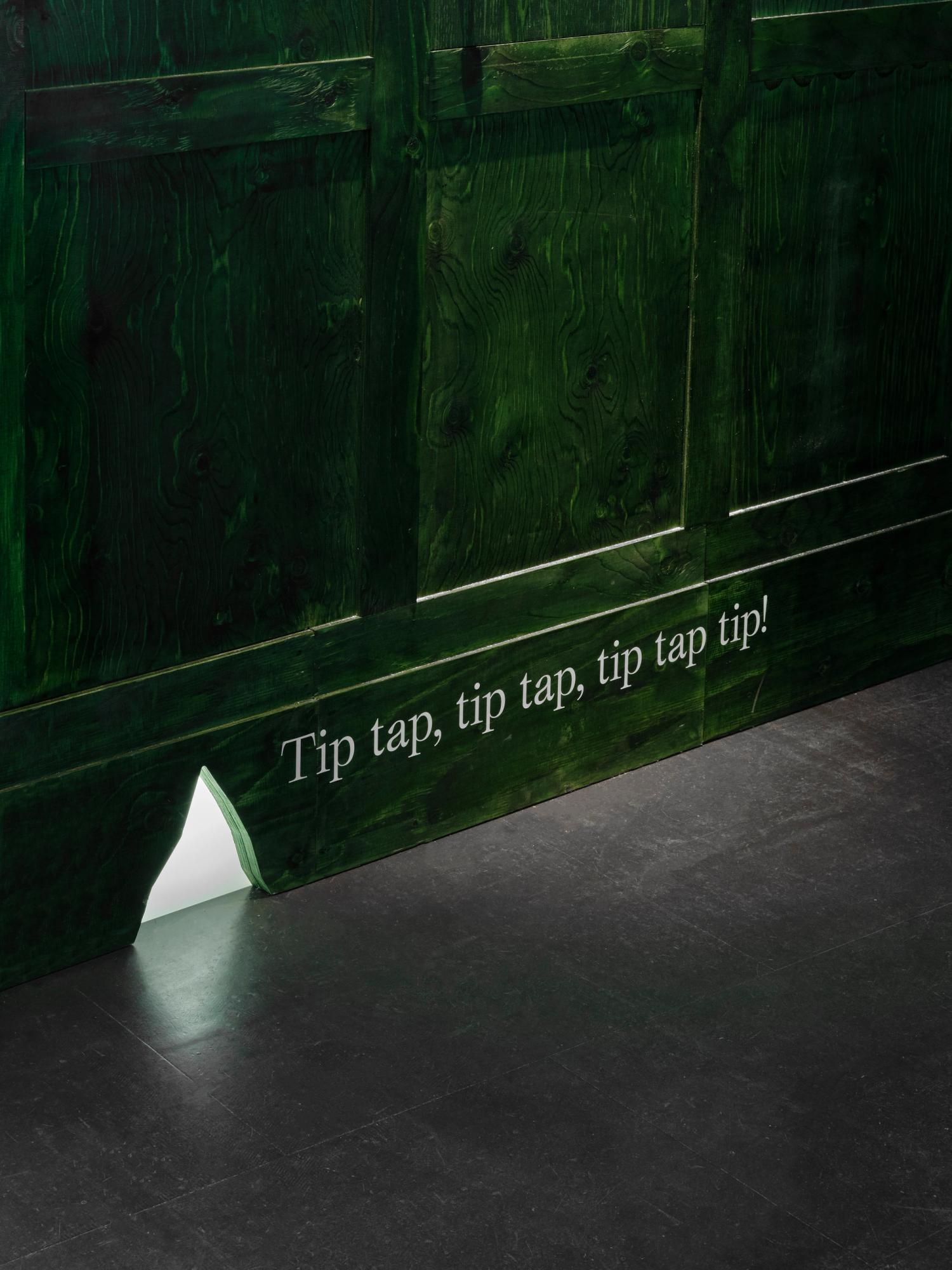 A close-up detail of a small jagged hole in the skirting of a dark green exhibition wall, with onomatopoeic text printed to the wall beside the hole