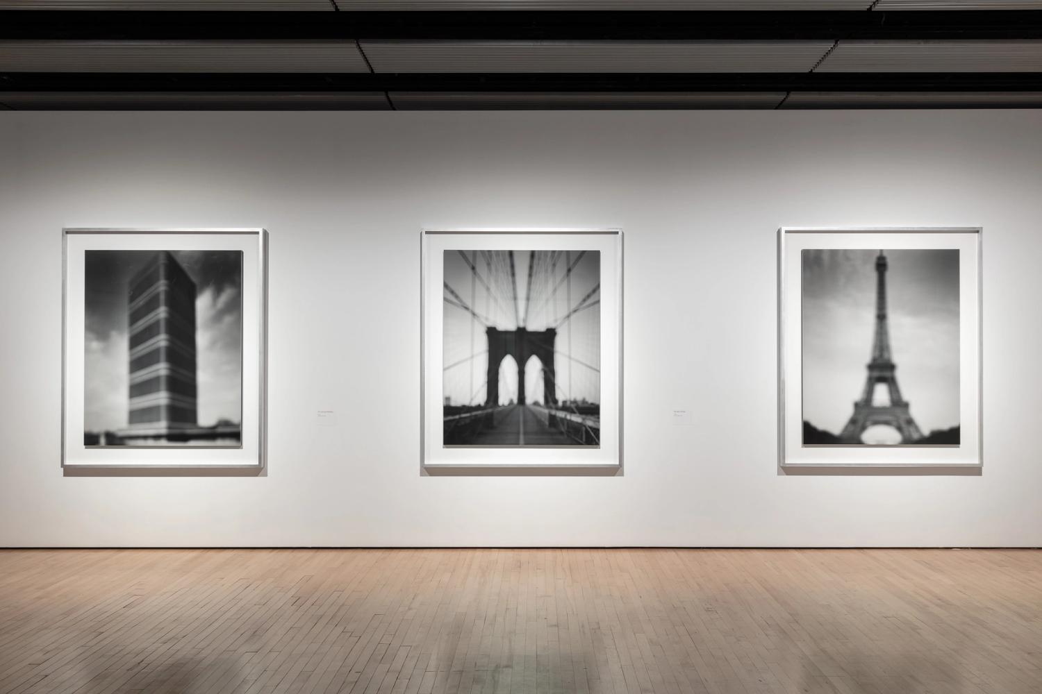 Photograph of a gallery wall with three framed black and white photographs, each showing a blurred image of an iconic landmark