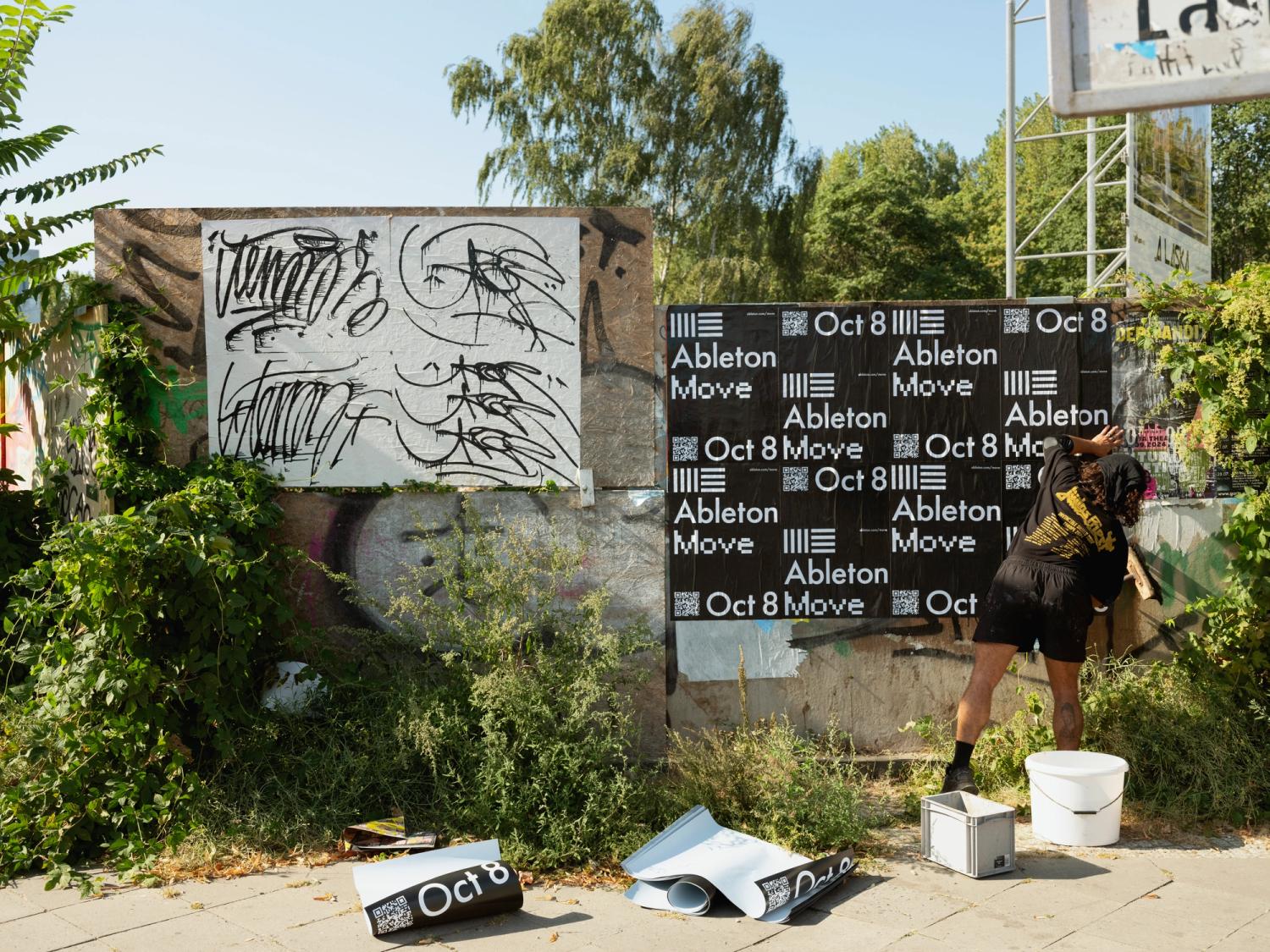 A street scene featuring a wall with graffiti and a series of black and white posters advertising a Ableton Move. A person is pasting the posters on the wall, surrounded by plants and urban decay elements.