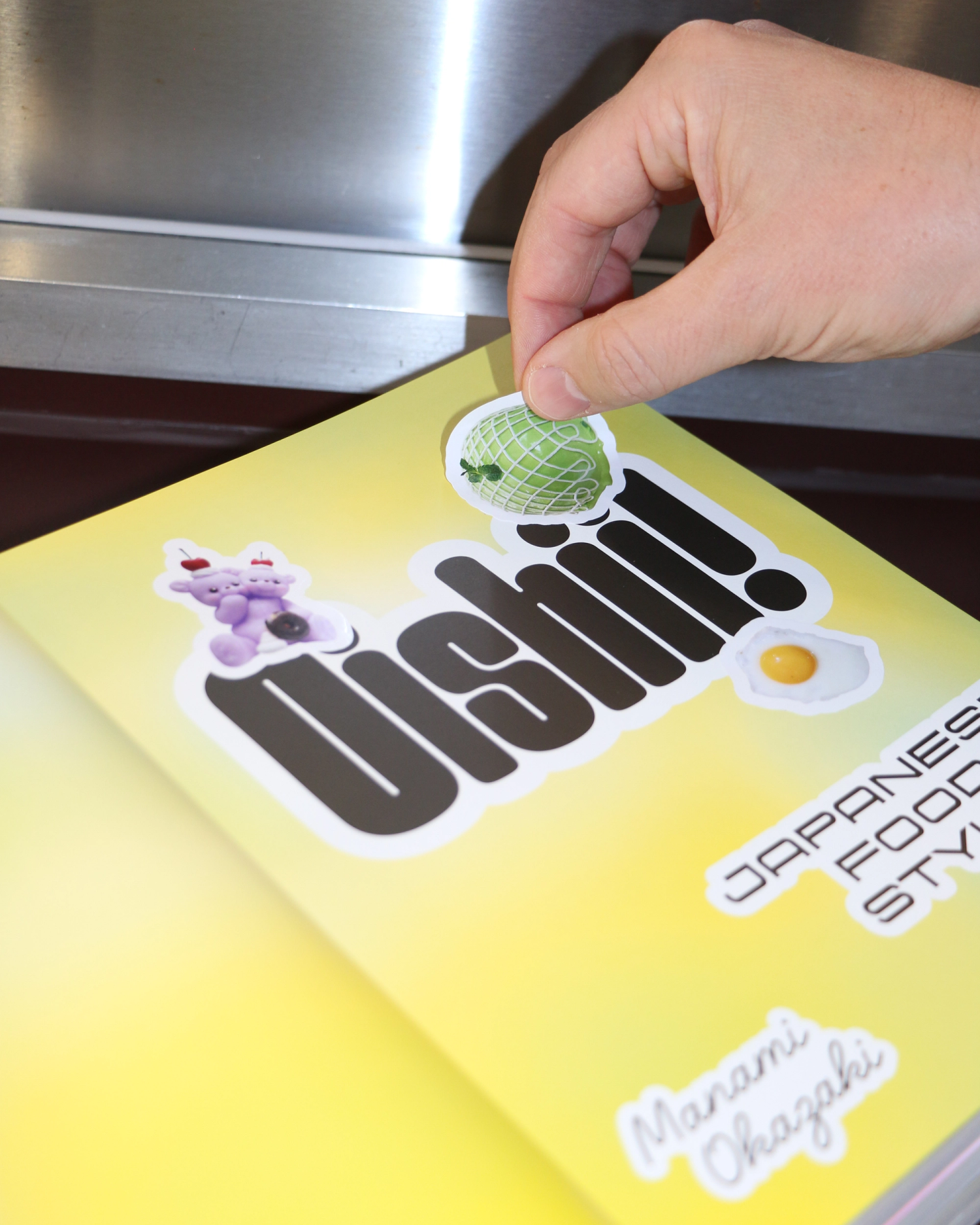 An open book with a yellow page background and the book title printed. A hand is placing a sticker of a green cake over the title.
