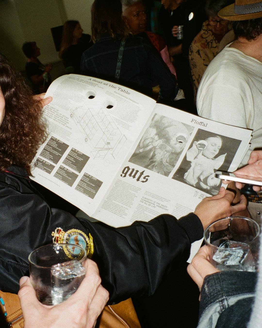 Person holding Ödd Paper, featuring two punched eye holes, surrounded by people with drinks at an event.