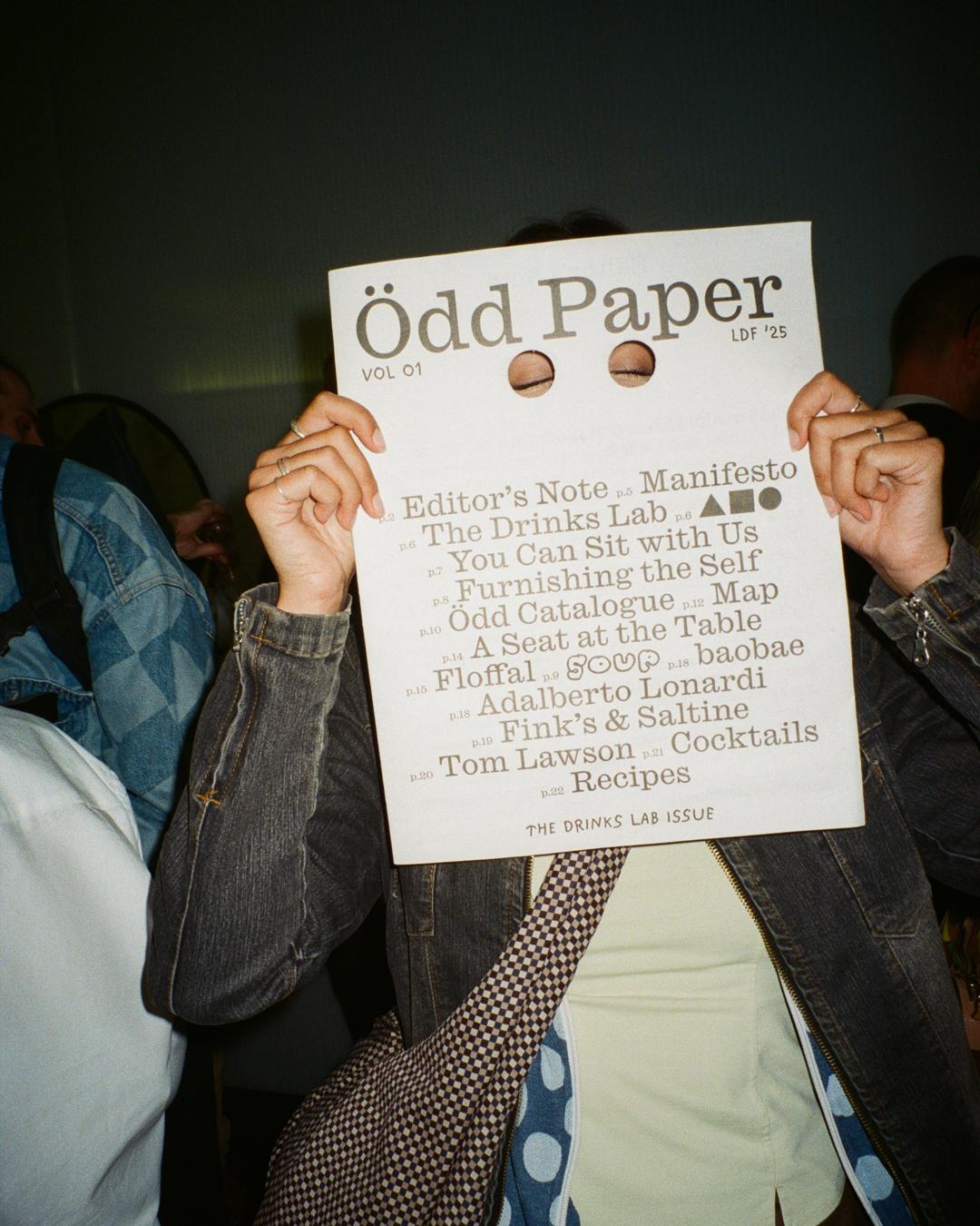 Person holding Ödd Paper against their face, with their closed eyes visible through the two holes cut into the newspaper. Text includes content list and “The Drinks Lab Issue.”