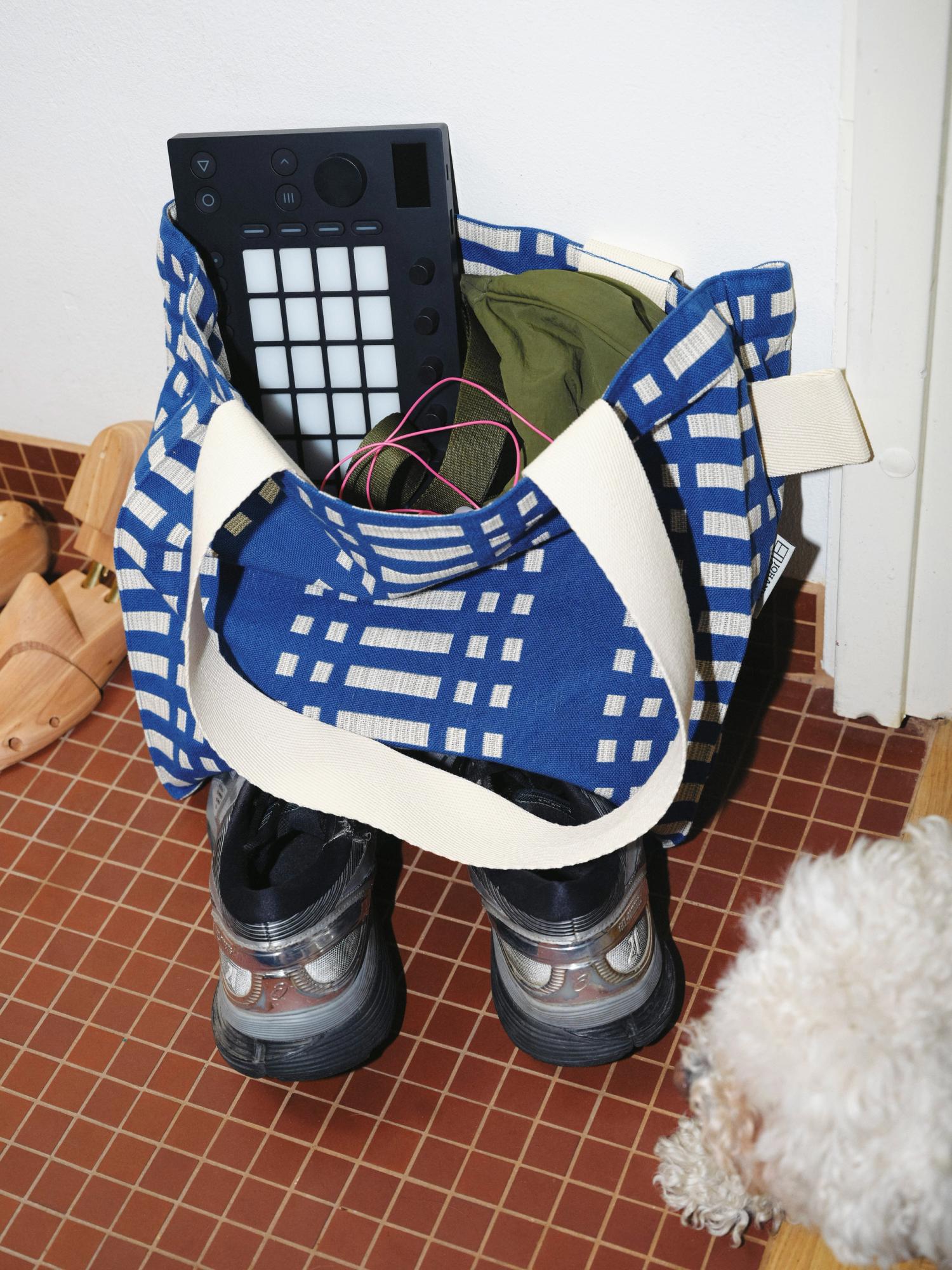 A blue patterned tote bag sits on a tiled floor, partially packed with items including Move. Below the bag, a pair of black and silver Asics sneakers are placed. In the bottom right corner, a white curly-haired dog is visible. The scene is indoors, with warm lighting.