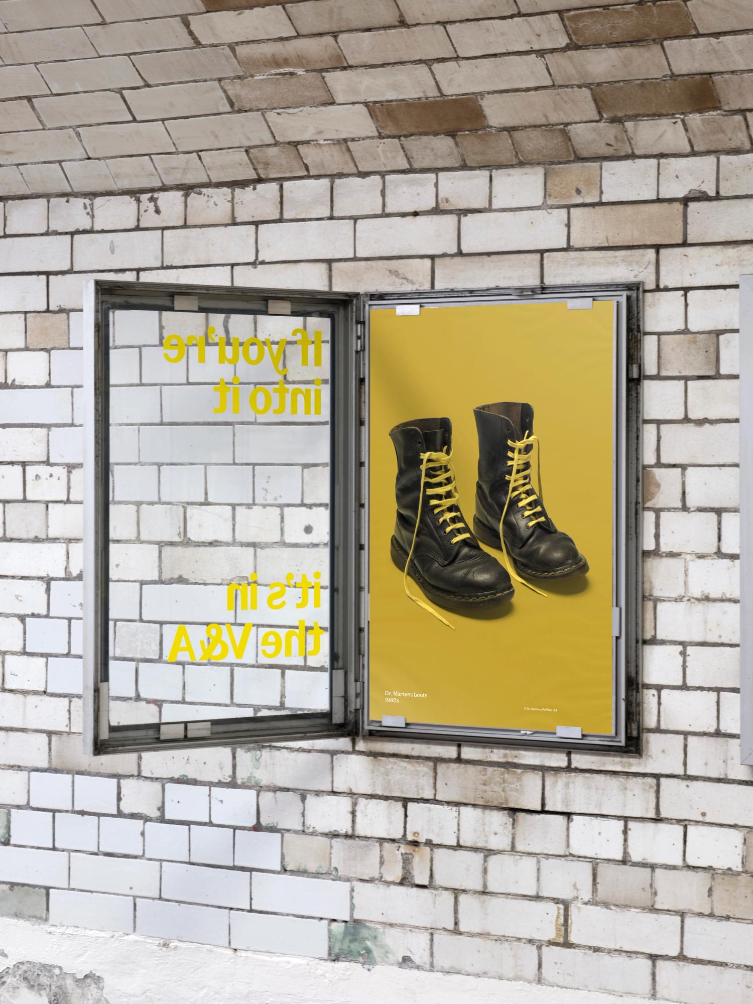 Photograph of an open poster display case on a brick wall, the poster within has a mustard yellow black background and an image of a pair of black boots with yellow laces. There is yellow writing on the outside of the poster glass.
