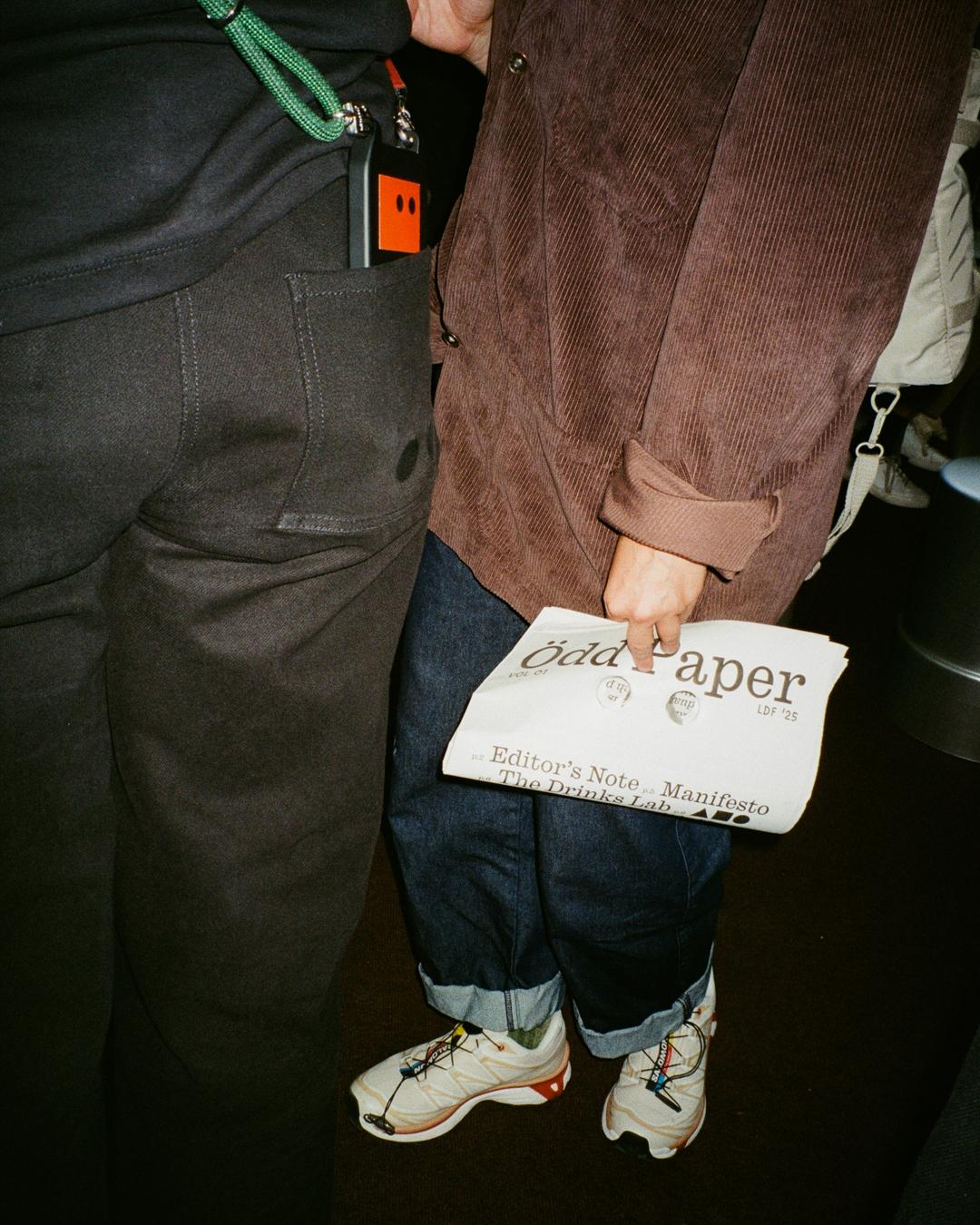 Person holds Ödd Paper with punched eye holes, dressed in a brown jacket and jeans, standing beside another person.
