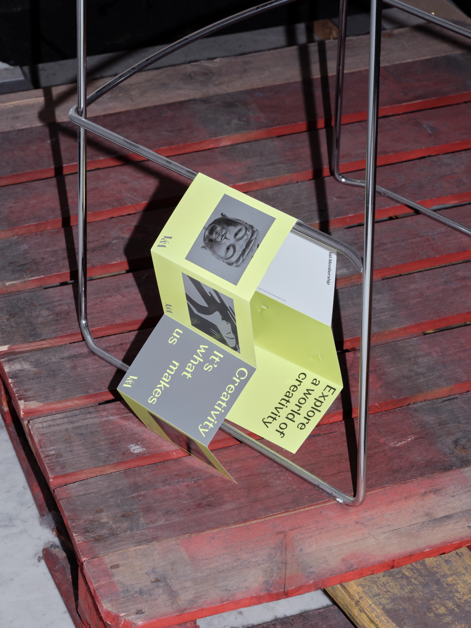 Photograph of a neon yellow and grey concertina leaflet draped over a stainless steel chair frame on a red pallet.