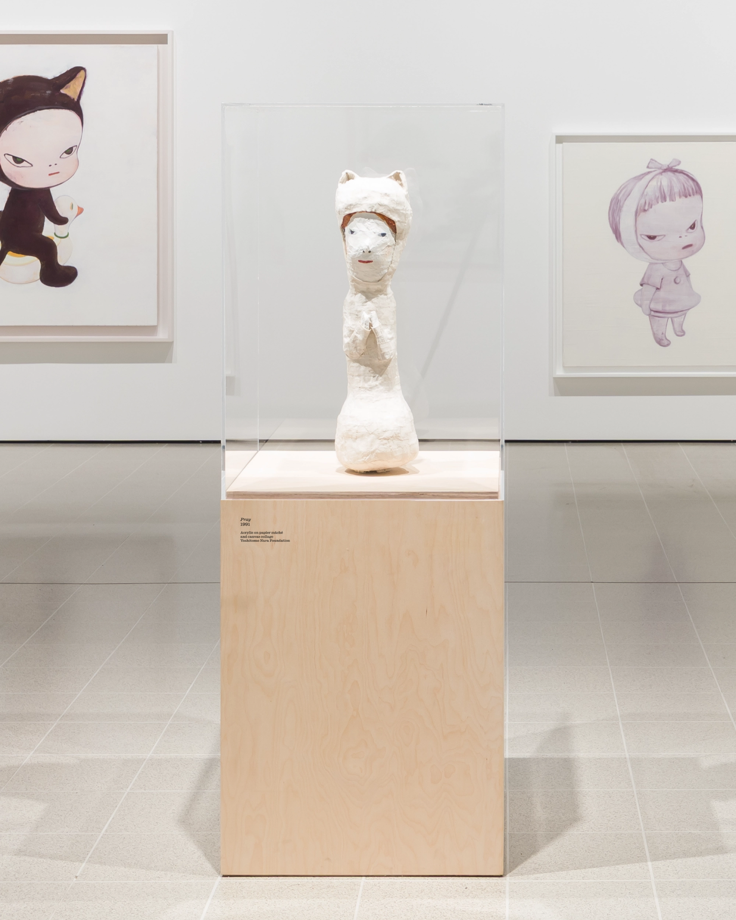 A wooden plinth sits centred in a gallery space, with two framed artworks on the wall behind. The plinth holds a white sculpture, and has a small caption printed to the corner.