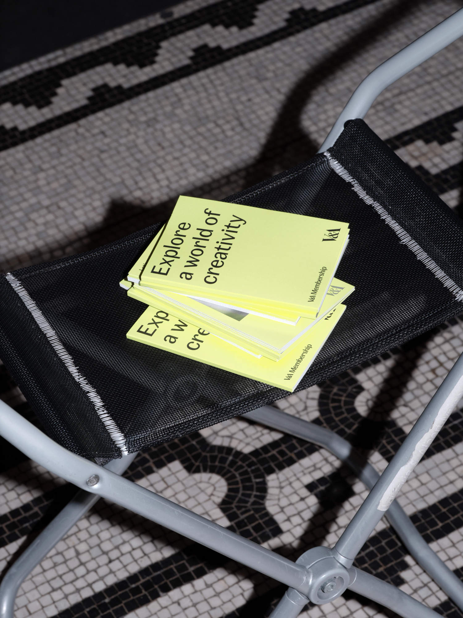 Photograph of a stack of neon yellow booklets on top of a fabric stool on a mosaic floor.