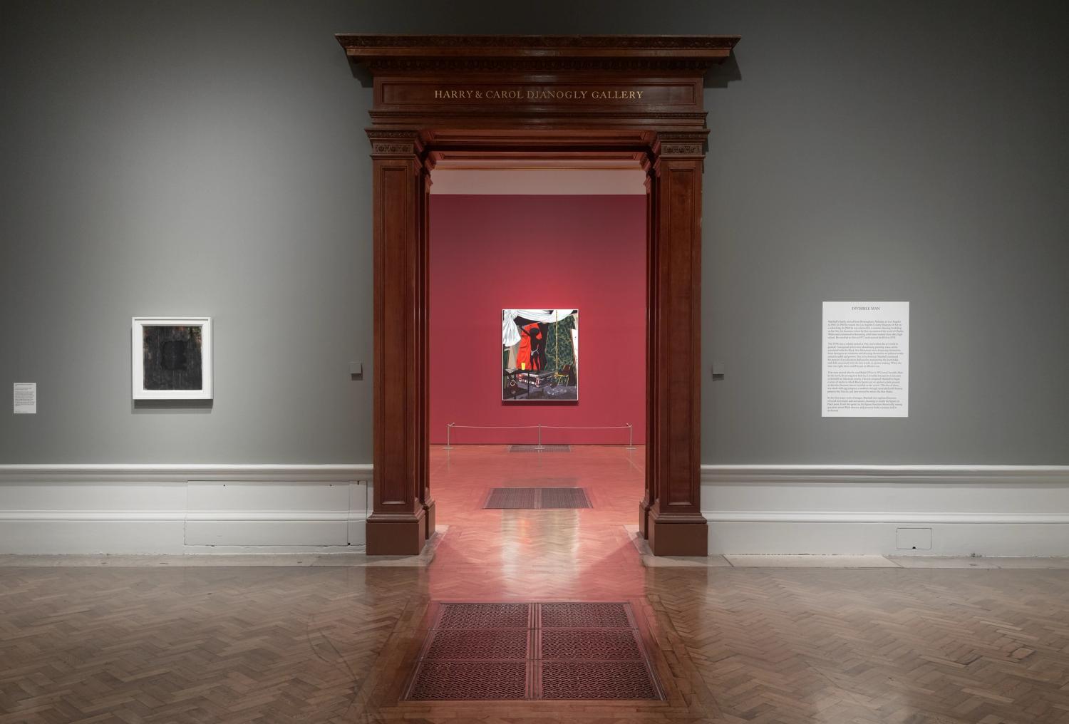 A framed artwork and two interpretation panels on a grey wall, flanking a grand wooden threshold. A second artwork is visible on a red wall framed in the doorway.