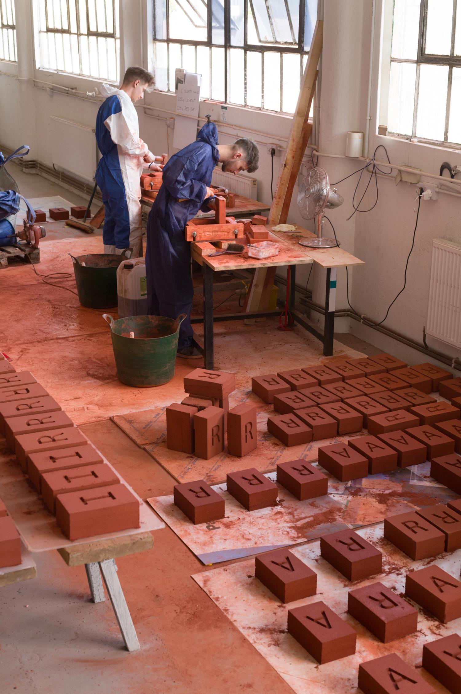 Two men in overalls stand at a workbench surrounded by red bricks