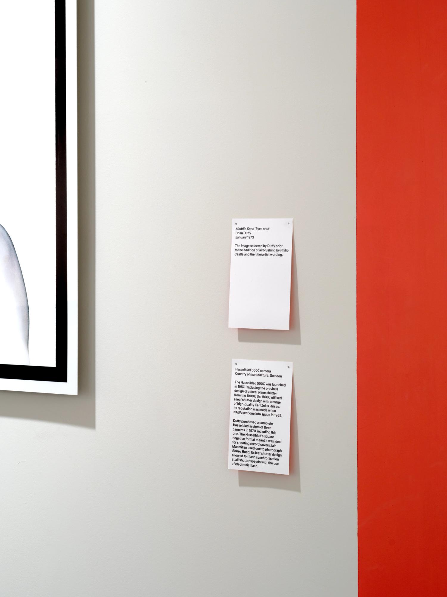 A photograph of two artwork captions pinned to a white gallery wall, with the red edge of the wall visible. The captions have black text printed on them and a red glow on the wall behind.