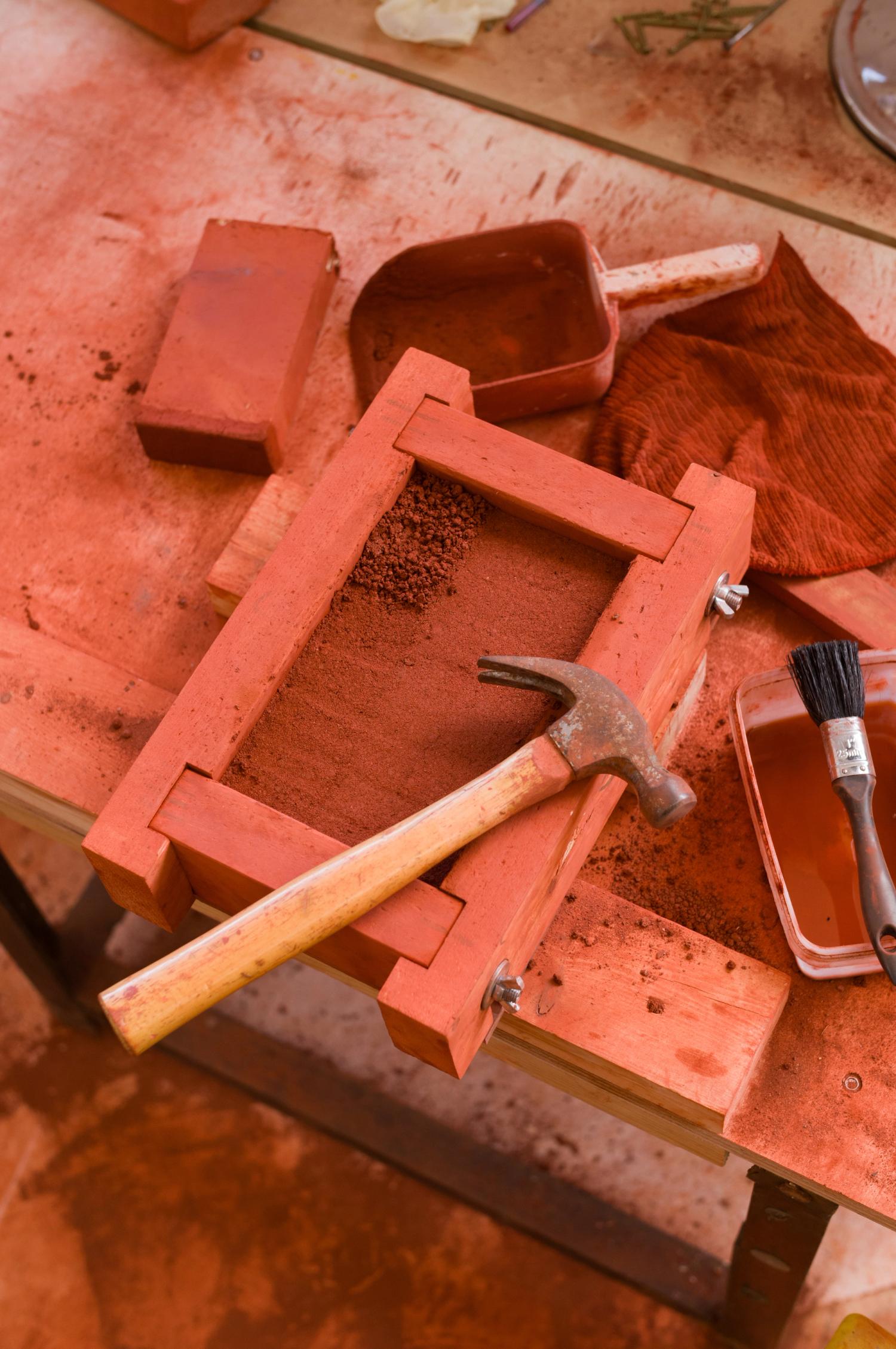 A workbench with red dust and tools and a wooden clamp frame filled with red powder with a hammer resting on top