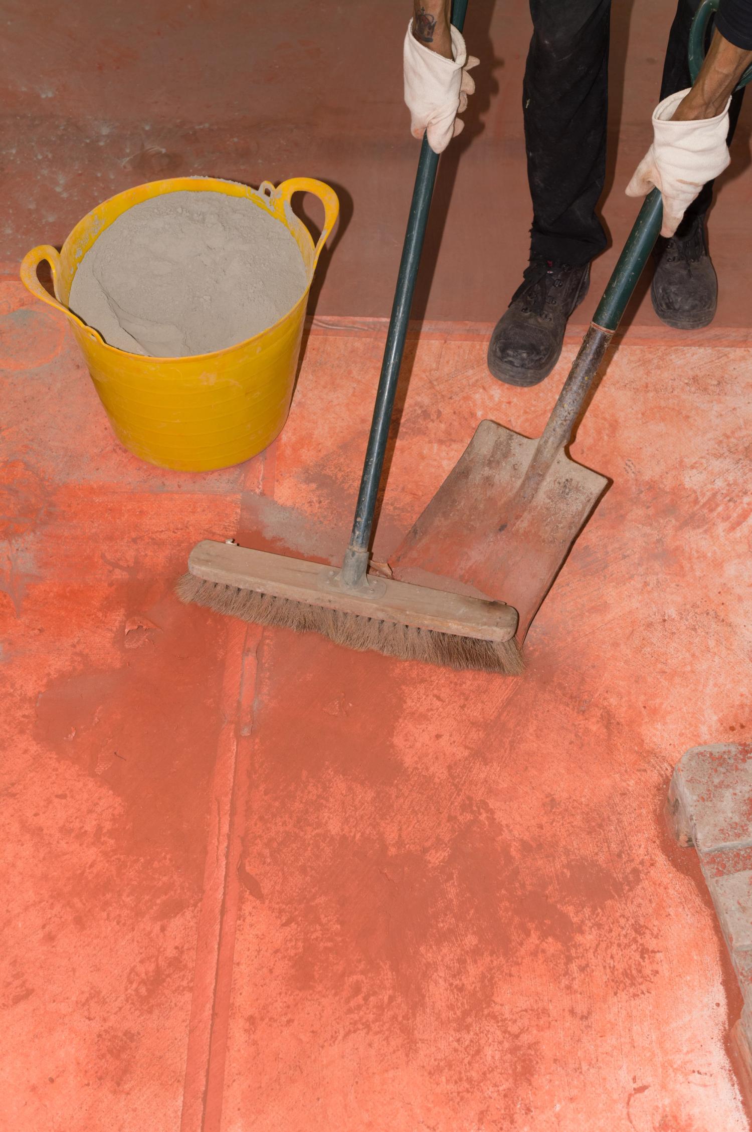 A person with black boots and white gloves sweeps up red dust next to a yellow bucket filled with grey powder